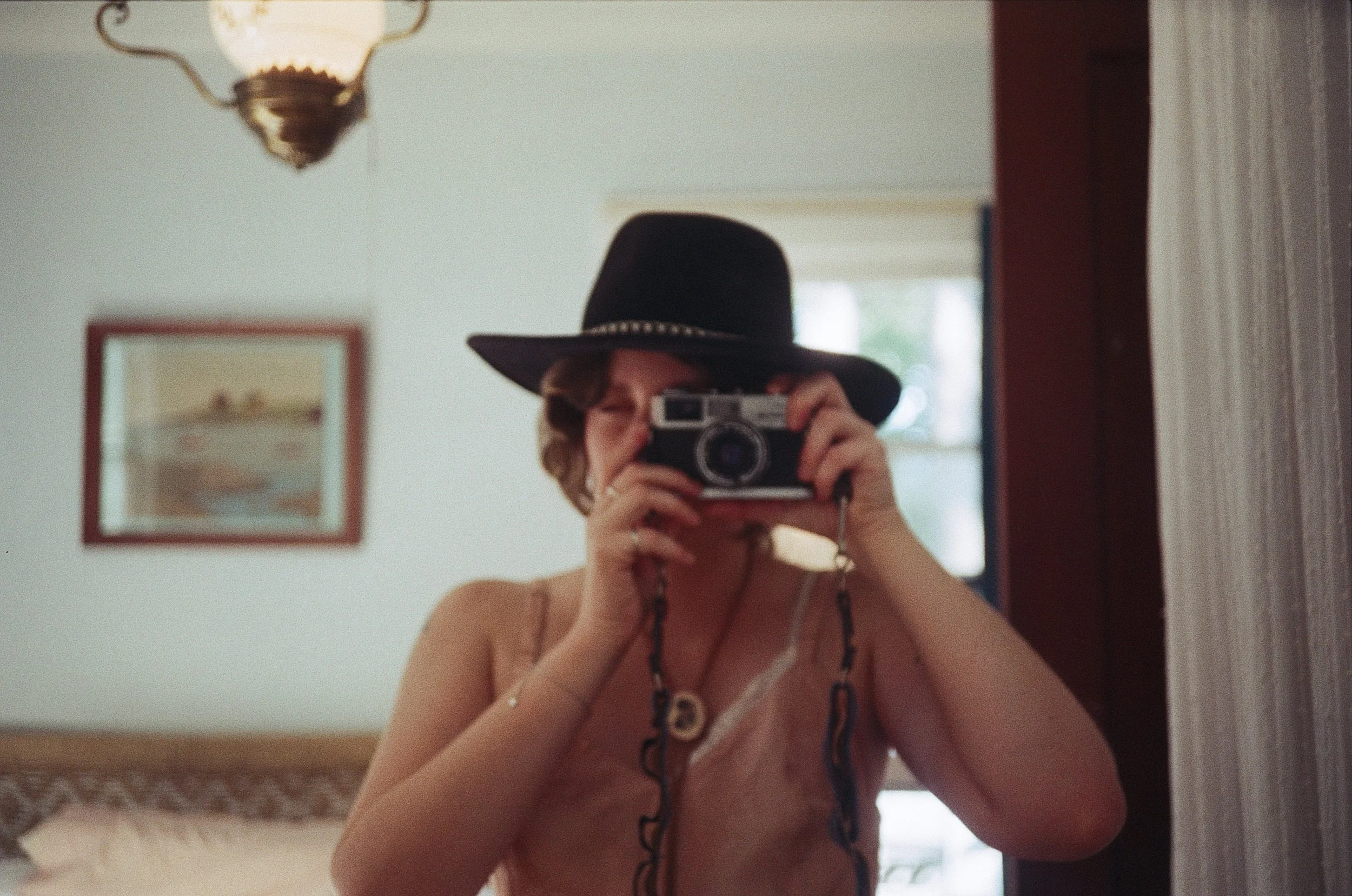 Person wearing a black wide-brimmed hat taking a photo with a vintage camera in a room with a hanging lamp and a framed picture on the wall.