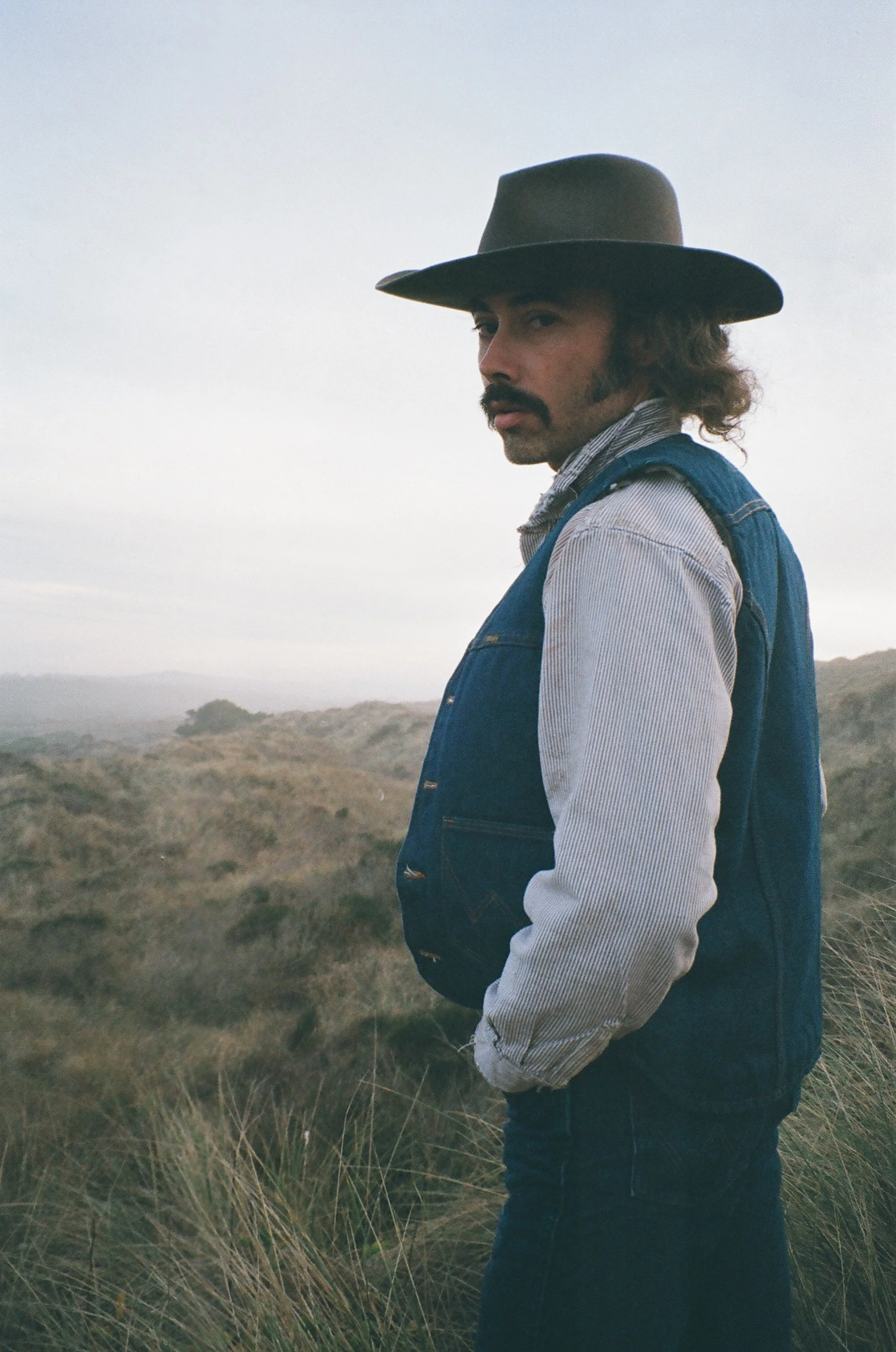 Man with a cowboy hat, mustache, and beard standing outdoors in a grassy landscape with cloudy sky.