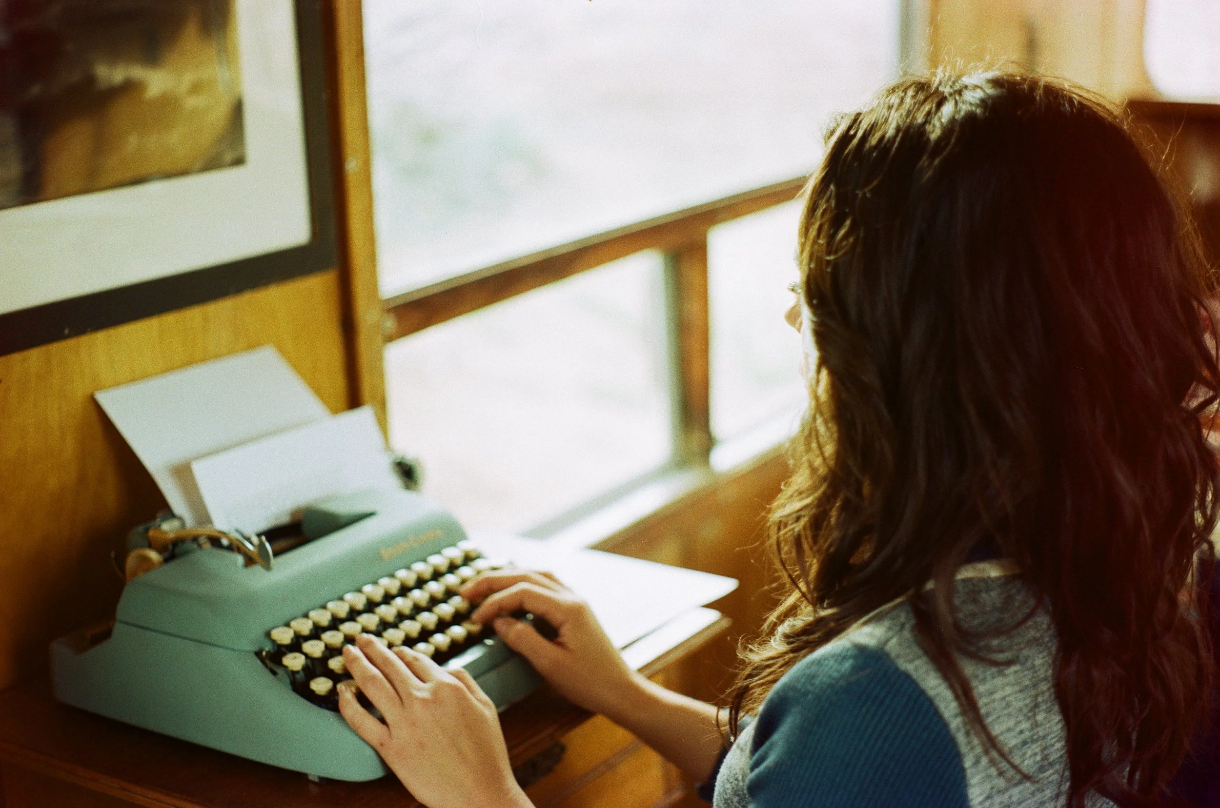 A woman with wavy brown hair, wearing a gray and blue sweater, is sitting at a wooden desk typing on a vintage light green typewriter, with papers inserted, next to a window.