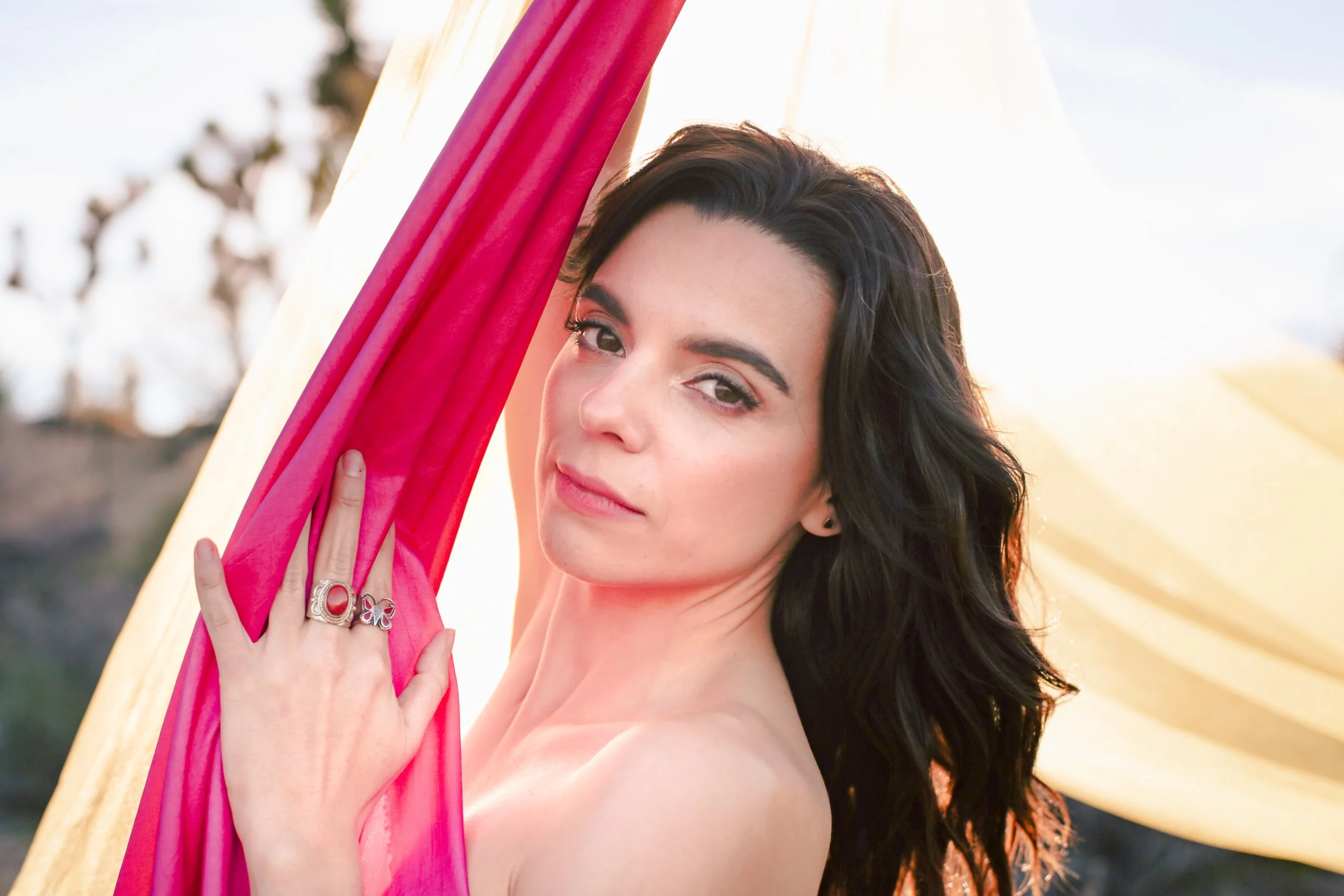 A woman with dark hair and light skin looking at the camera, holding pink and yellow fabric outdoors.