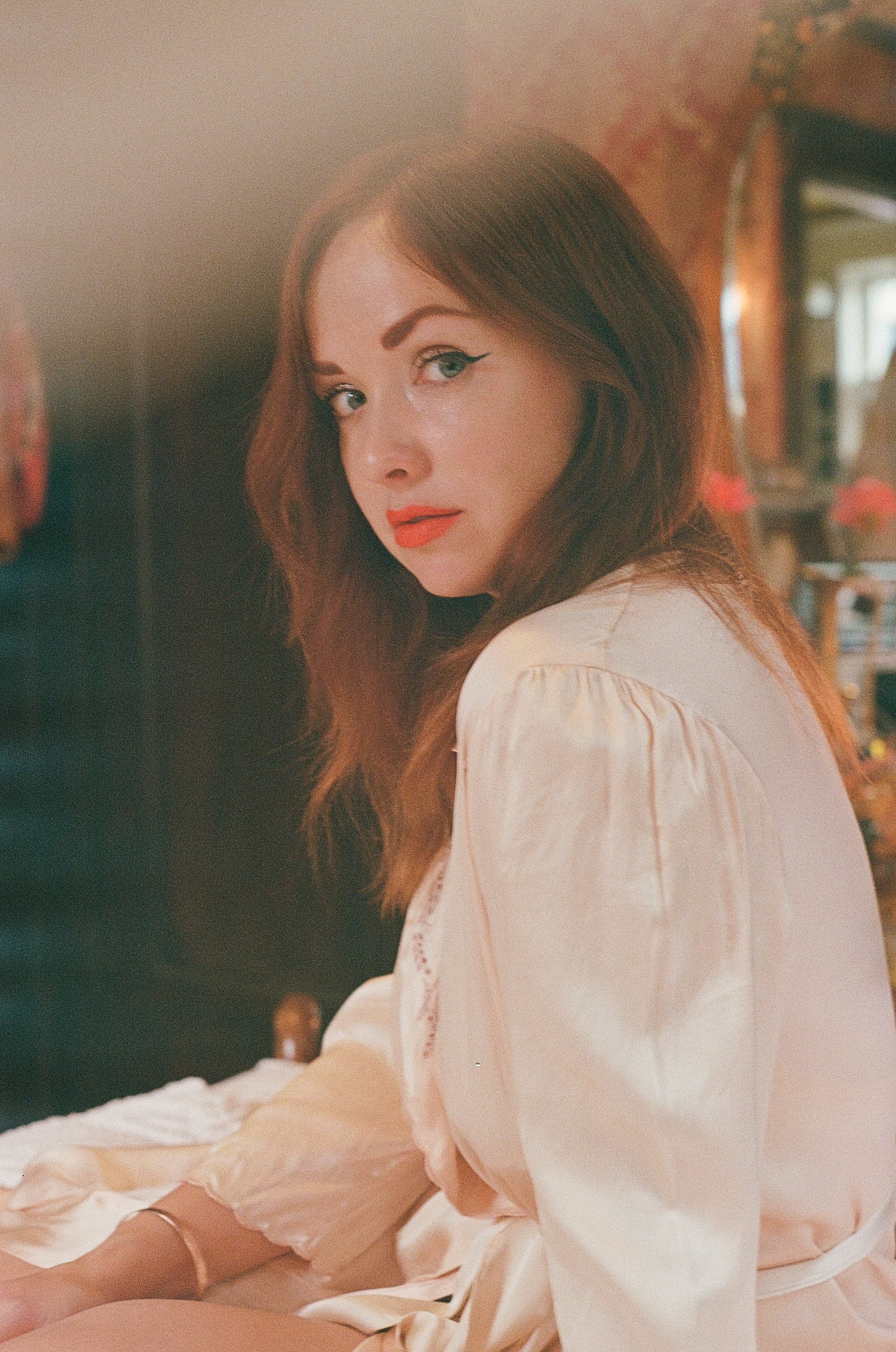 A woman with red hair, makeup, and a beige satin robe sitting indoors in a warmly lit room.