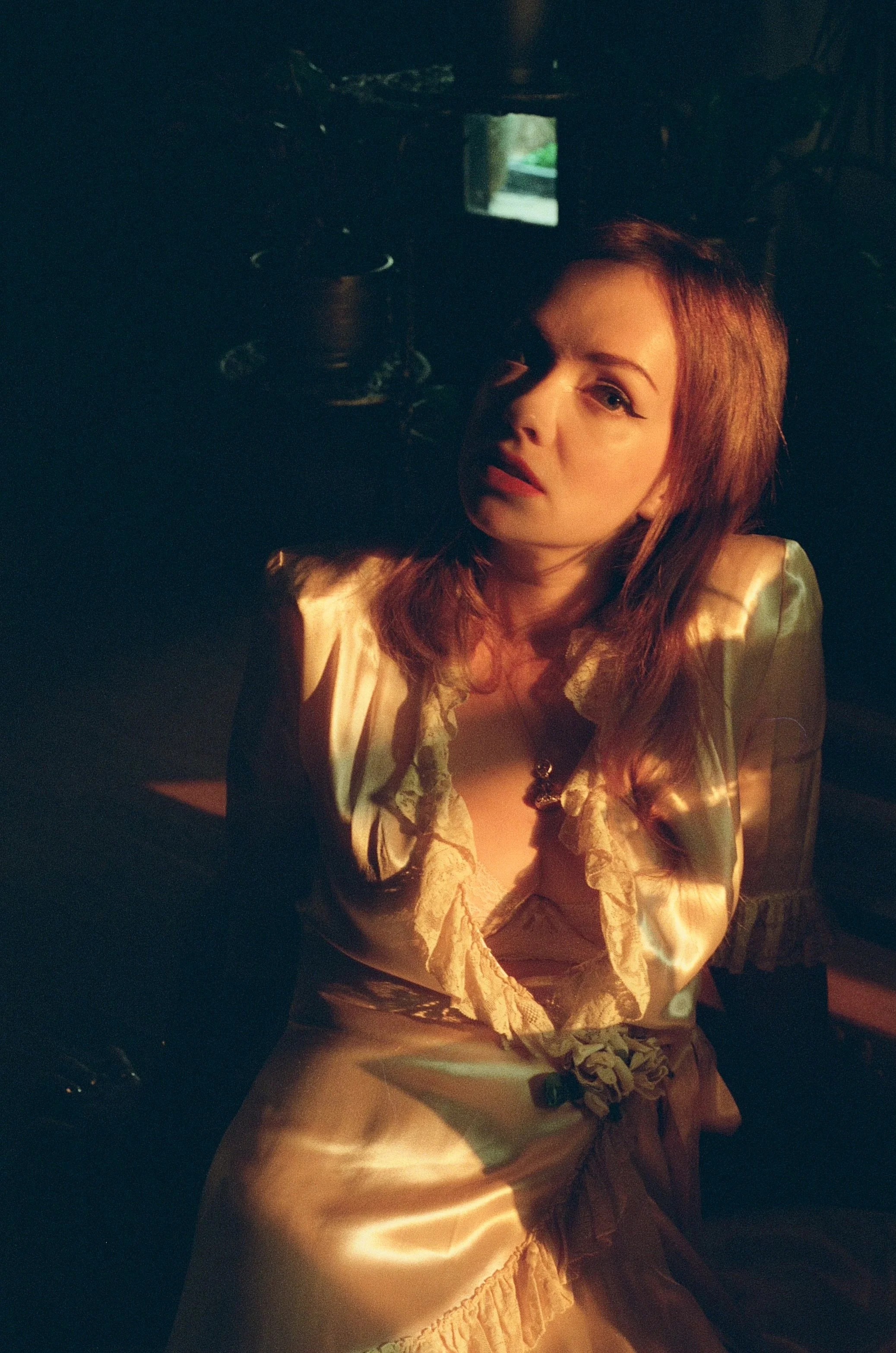 A woman with red hair and winged eyeliner wearing a cream satin dress with lace details, sitting in a dimly lit room with a small window behind her.