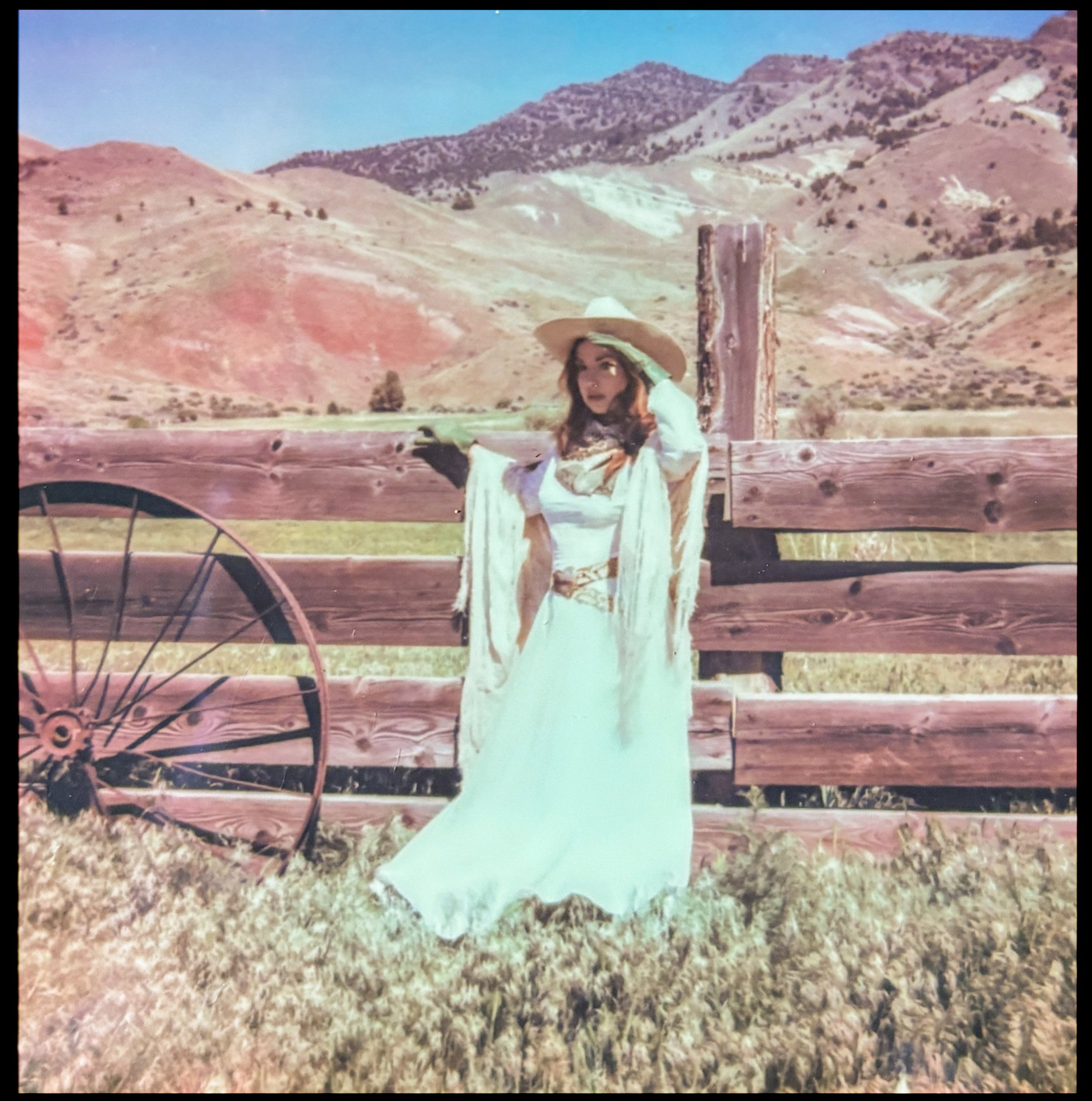 A woman dressed as a cowboy wearing a white dress and a wide-brimmed hat, standing in front of a wooden fence with mountains in the background.