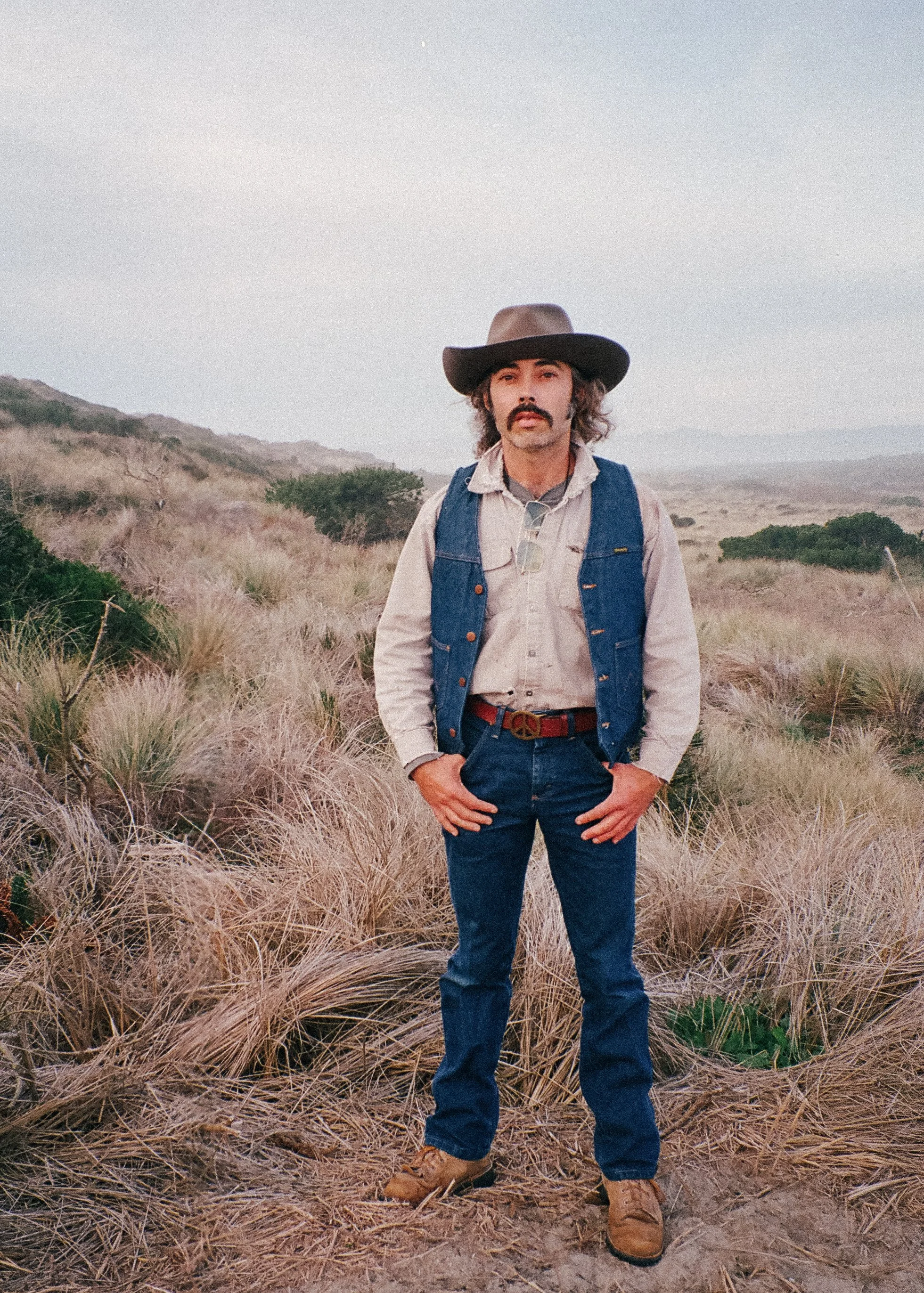 A man dressed in vintage cowboy attire with a wide-brimmed hat, denim vest, button-up shirt, jeans, and boots standing in a grassy, hilly landscape during daytime.