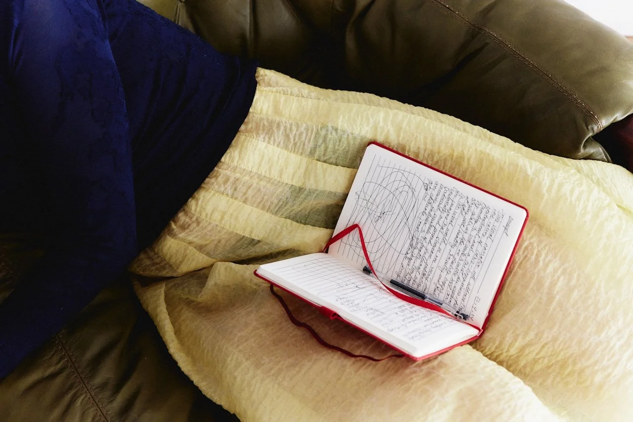 An open journal with handwritten notes and sketches resting on a beige cushion, with part of a person's blue sleeve and brown leather couch visible.
