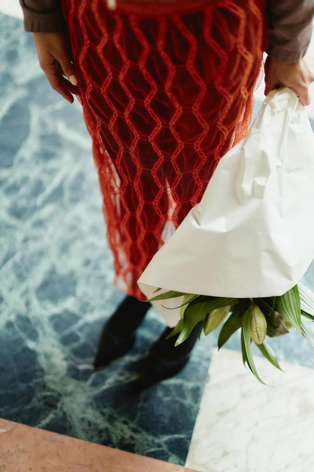 Person holding a bouquet of flowers wrapped in white paper, wearing a red lace dress and black boots, standing on a marble floor.