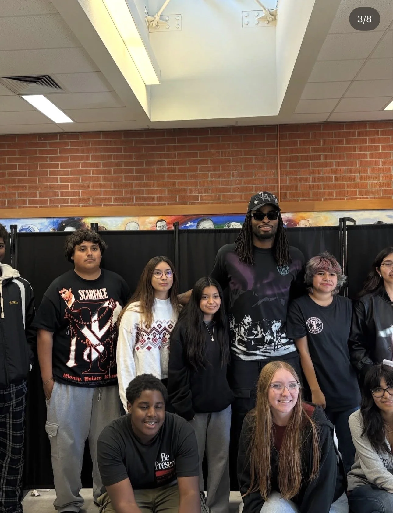 A group of diverse young students and a tall man with dreadlocks and sunglasses standing together in a school hallway, with a brick wall and a black divider behind them.