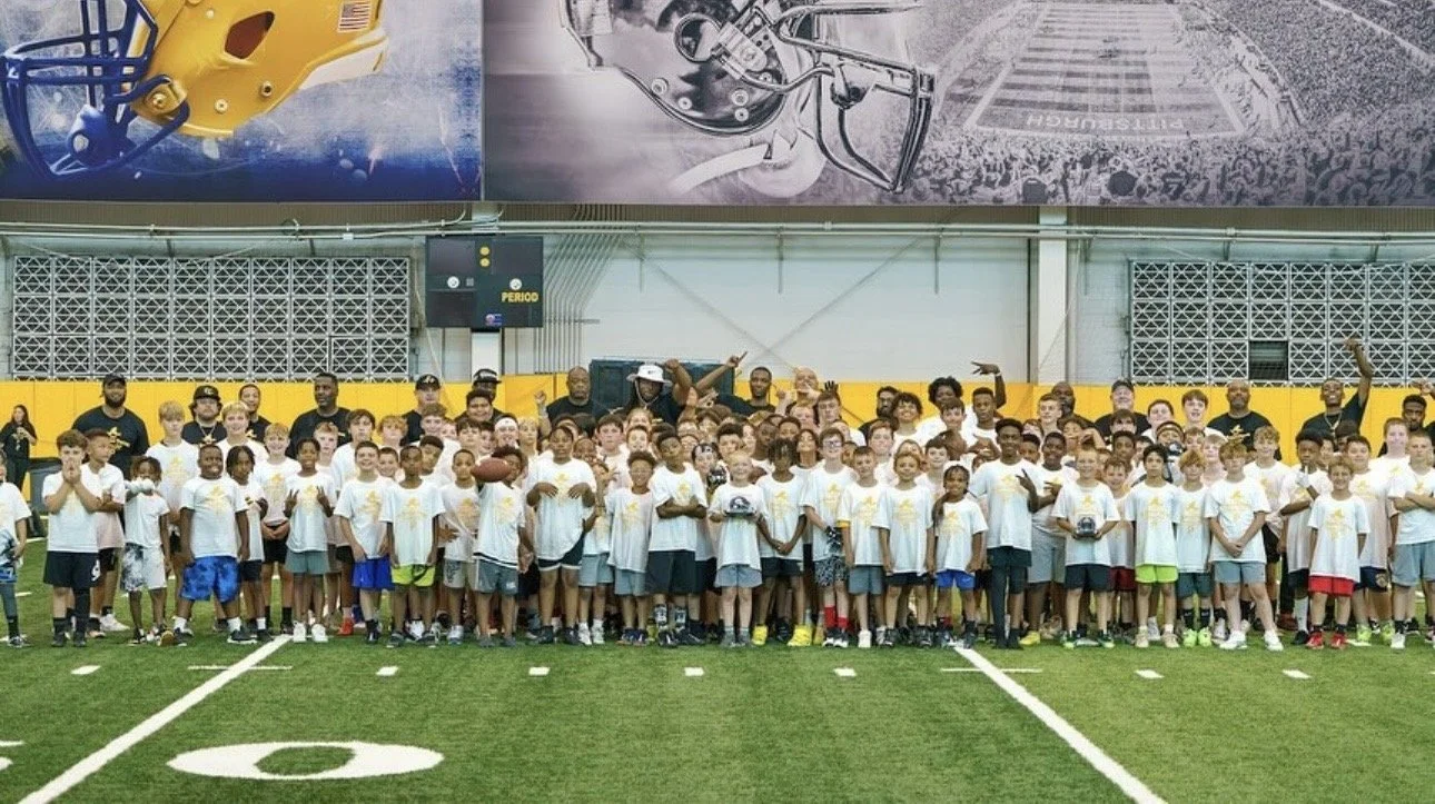 Group of children and coaches on an indoor football field, wearing matching white T-shirts, with large banners of football helmets hanging above the group.