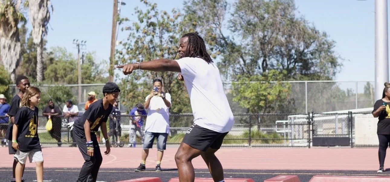 A man with long dreadlocks is smiling and encouraging children during an outdoor activity on a track field. The children are in black shirts, and one is smiling and bending forward. There are other people in the background, some taking photos.