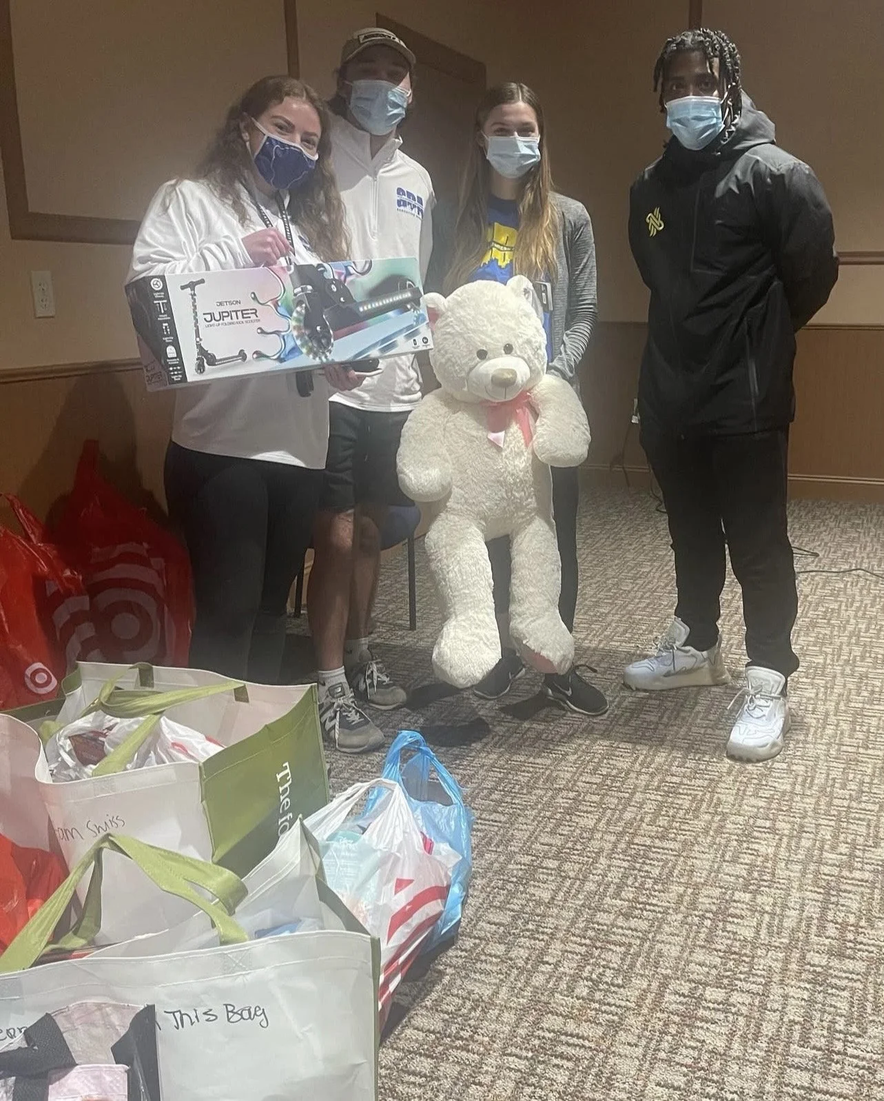 Four people wearing face masks standing with a large teddy bear, a box of a scooter, and several bags on the floor, in an indoor setting.