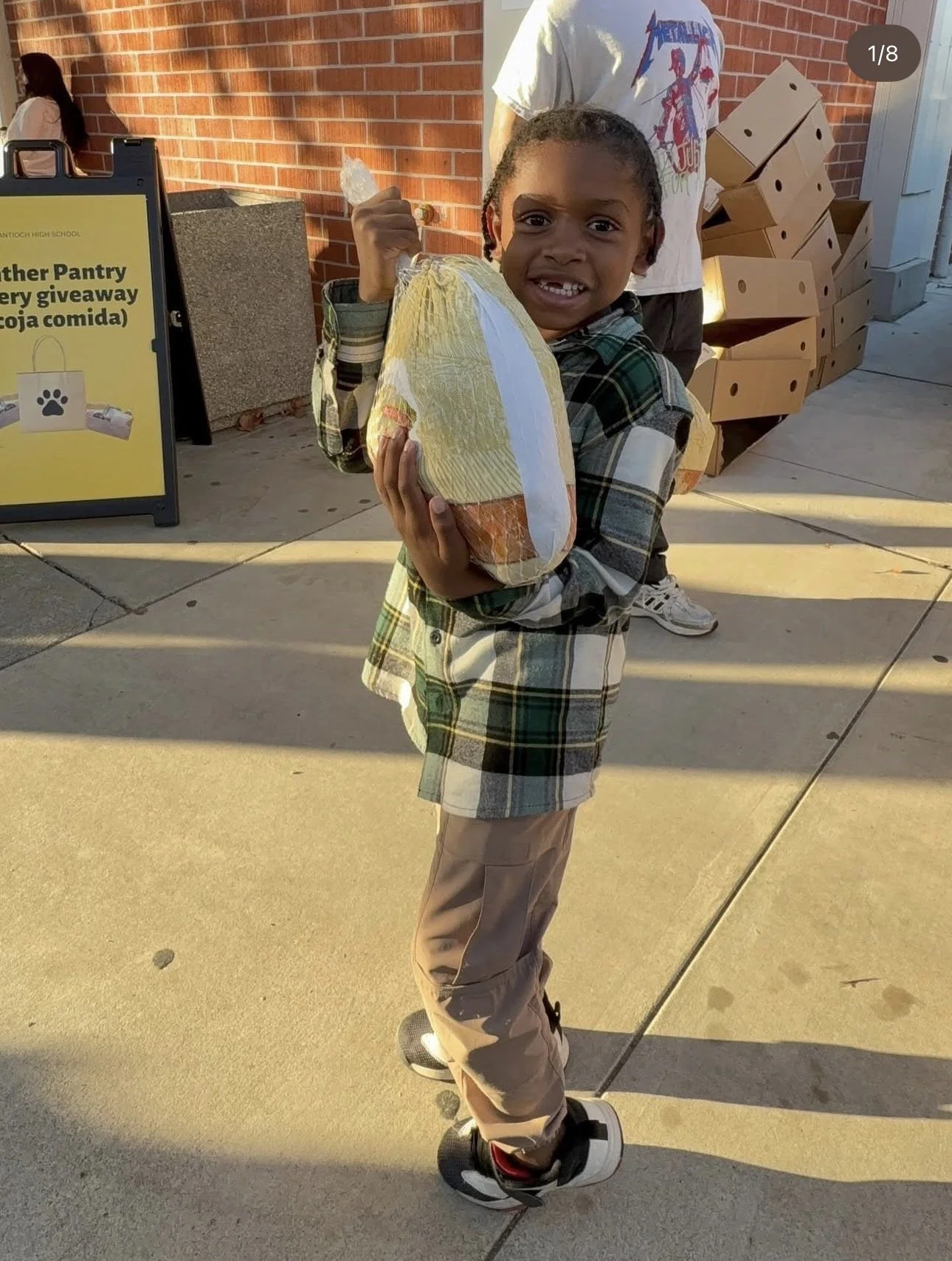 A young boy smiling and holding a large bag of food, standing outside near a brick wall and a donation sign for a pantry. The boy is wearing a plaid shirt and is on a hoverboard.