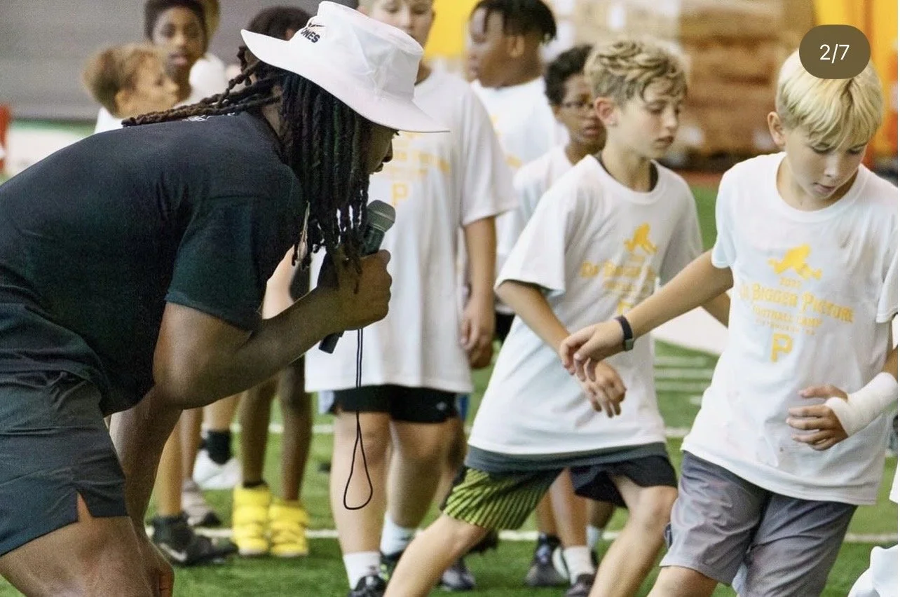 An adult coach or trainer with long dreadlocks, wearing a white hat and black shirt, holding a microphone, coaching a group of children in a sports drill indoors. The children are wearing matching white T-shirts and are participating actively in the 