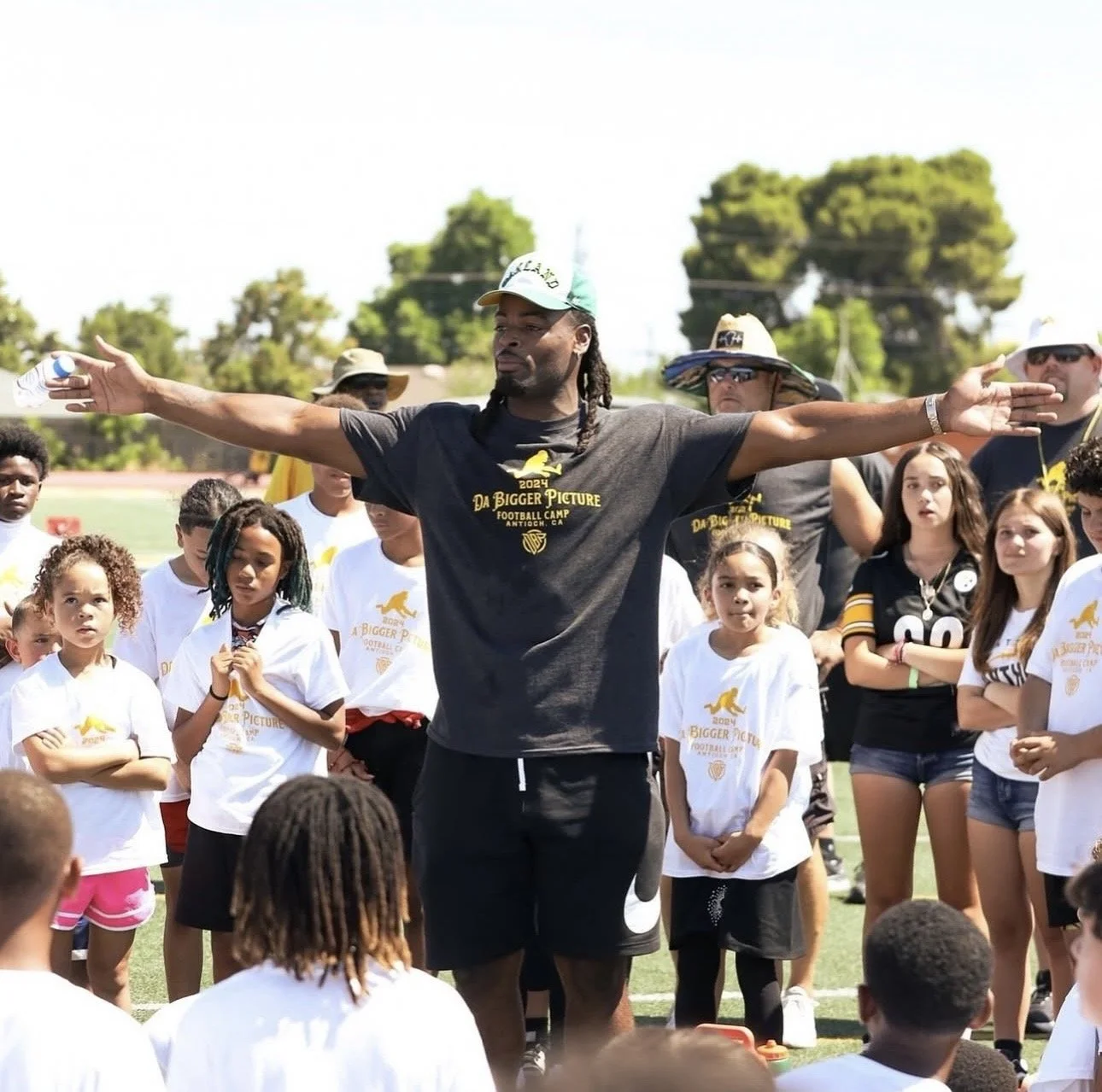 A football coach with outstretched arms standing in front of children and other adults during a football camp on a field.
