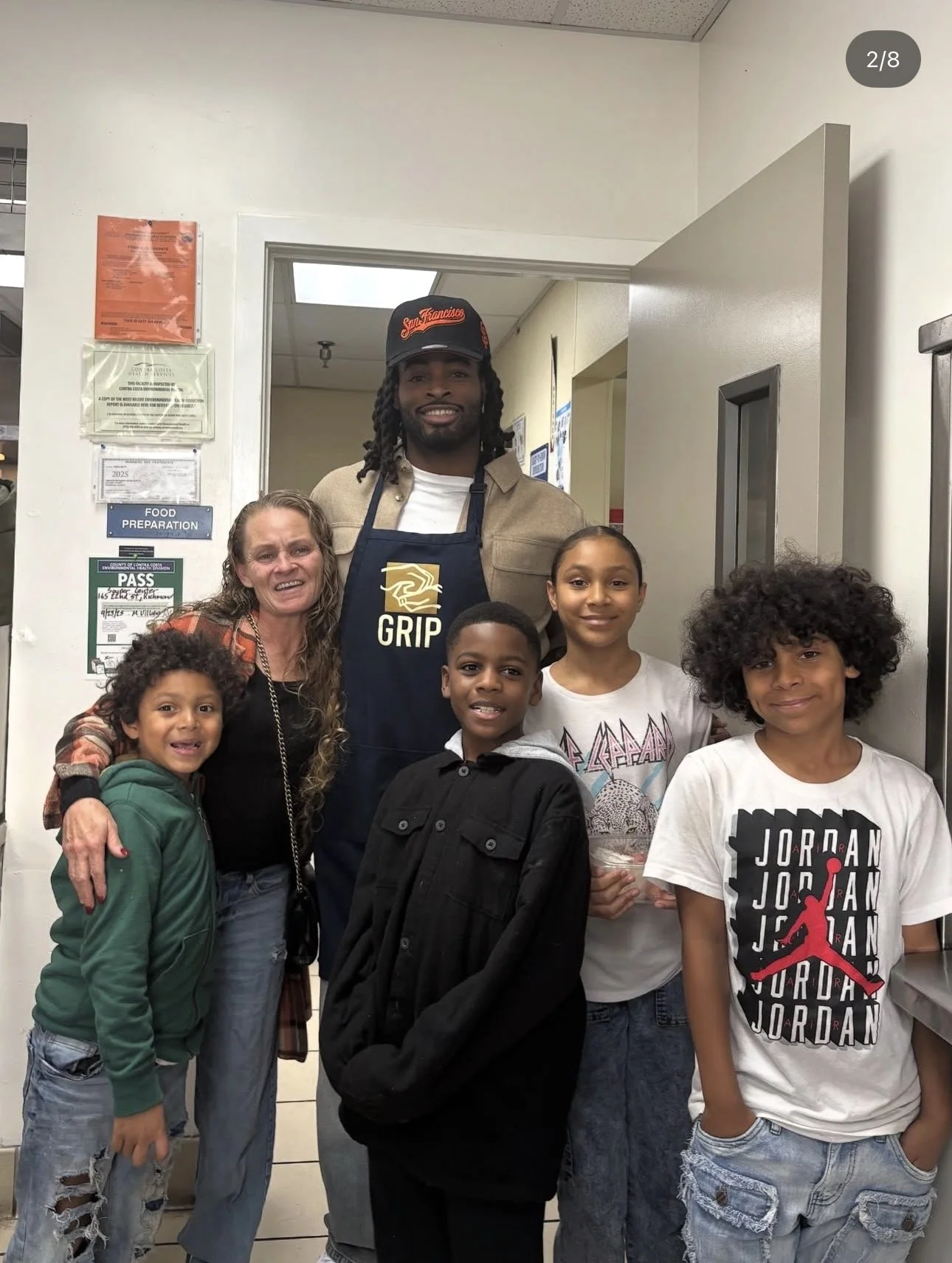 Group of people inside a room, including a man wearing a cap and apron with a Grip logo, and five children standing around him.