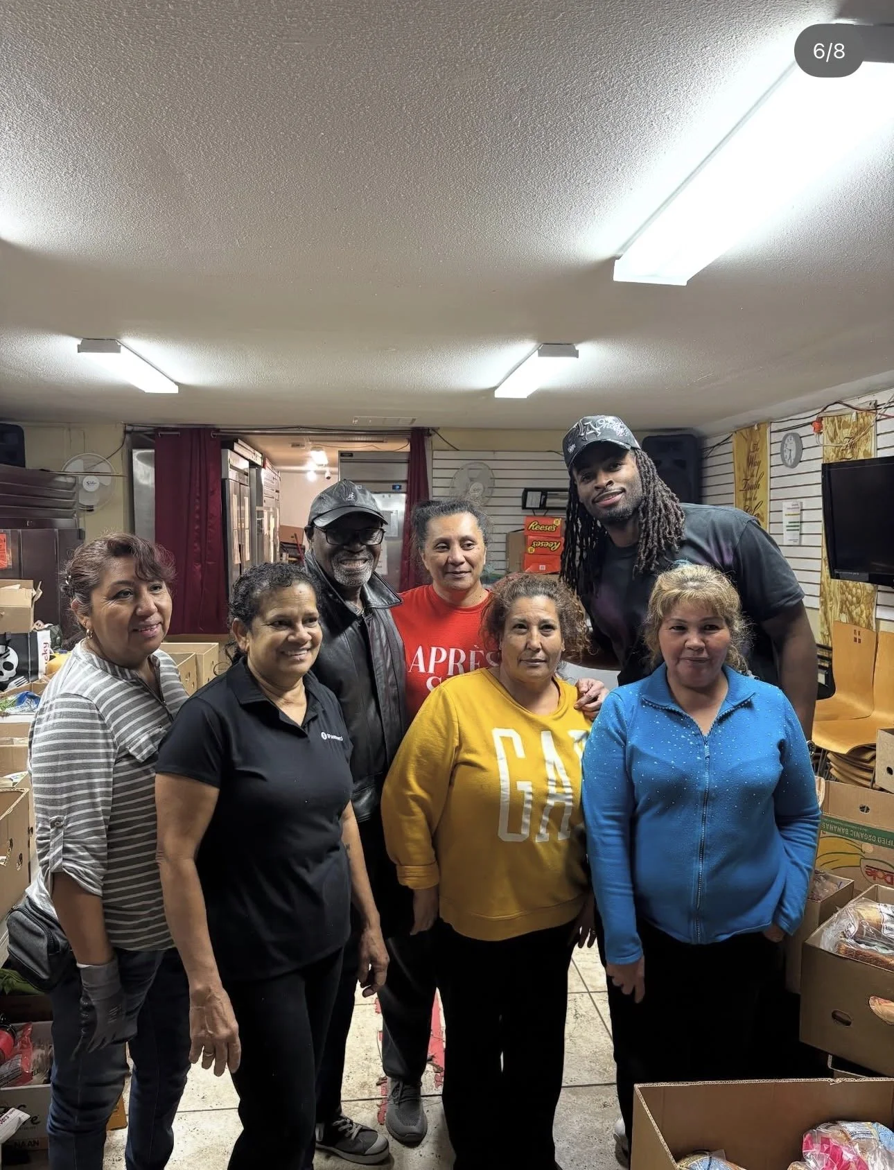 Group of seven diverse people standing together indoors, smiling for a photo, with boxes and shelves in the background.
