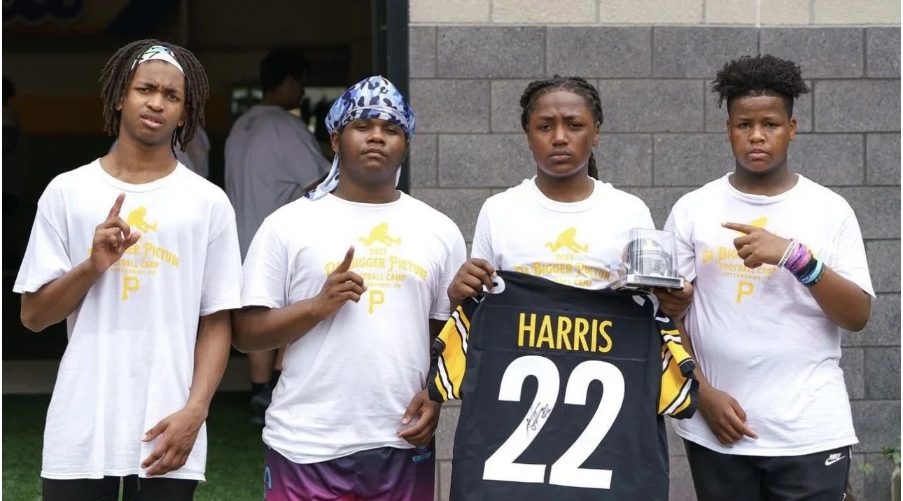 Four young girls standing in a row, wearing white t-shirts with yellow graphics, and making a gesture with their index fingers. One girl holds a black sports jersey with the name 'HARRIS' and the number '22' on it, along with a clear plastic box. The