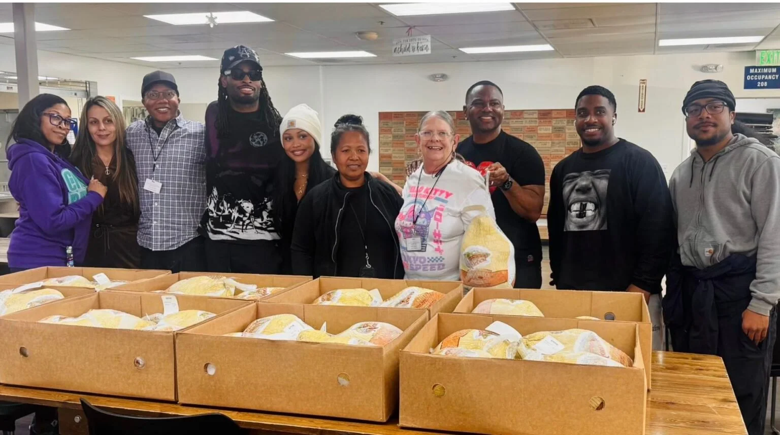 Group of ten diverse people standing behind boxes of packaged food in a room with white walls, some smiling at the camera.
