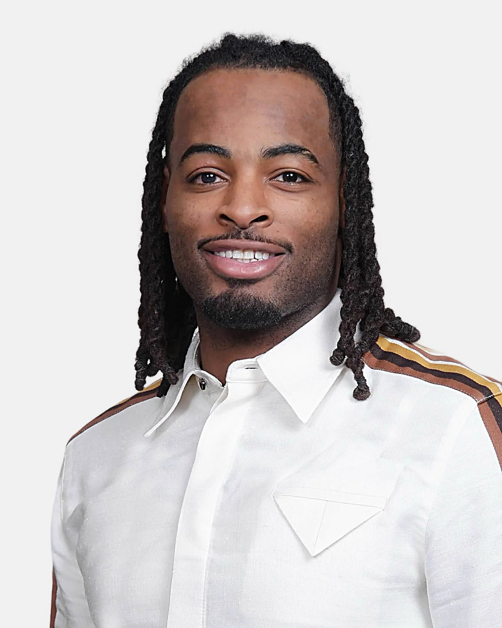Portrait of a smiling man with long curly hair, wearing a white shirt with patches of brown, black, and yellow on the shoulders, against a plain white background.