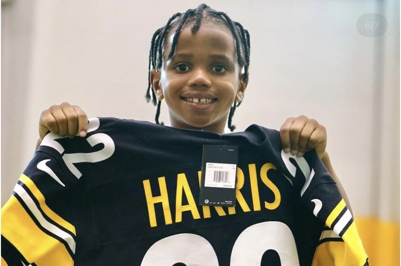 Young boy with braided hair smiling and holding up a football jersey that has the name Harris and the number 20.