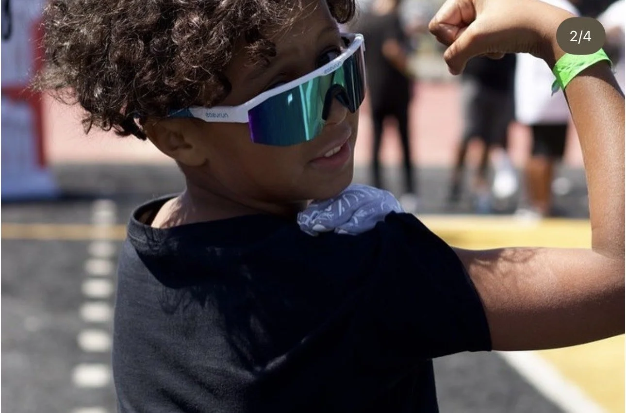 A young child with curly hair wearing reflective sunglasses and a black shirt, showing off their flexed arm muscle at an outdoor event.