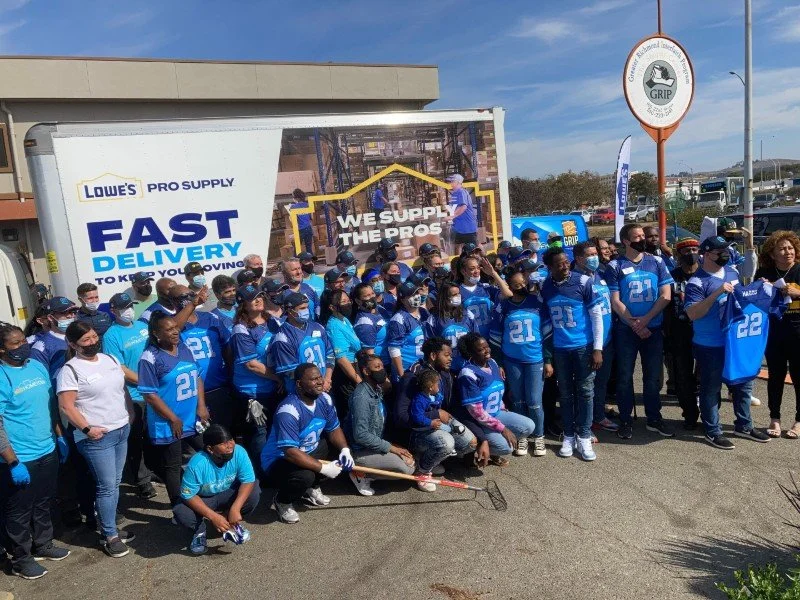 Group of people wearing blue jerseys and masks gathered outside a retail store, holding brooms and a shovel, with a big truck and promotional signs in the background.