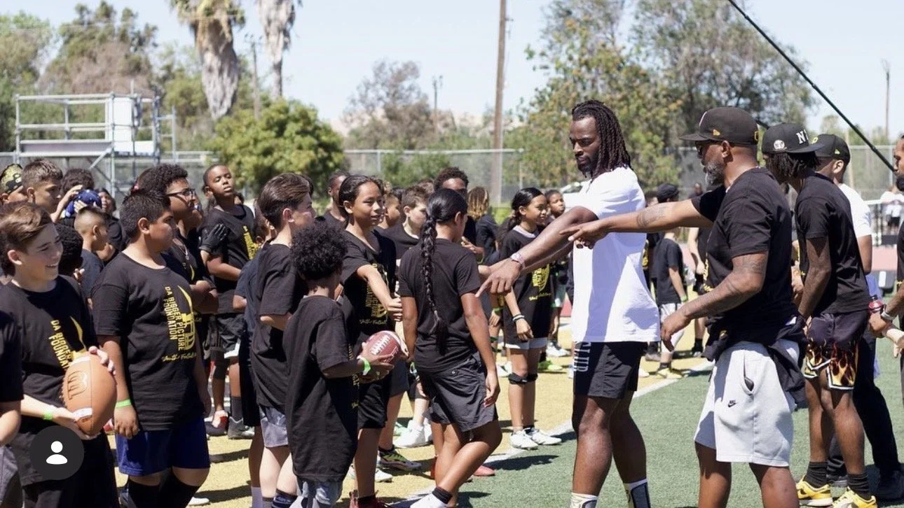 Group of children and adults on a football field during a youth football camp or clinic, with two adults instructing or coaching the children.