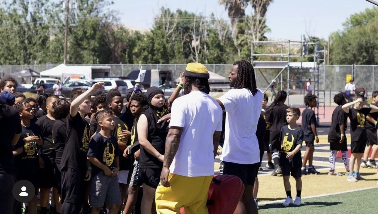 Group of children and teenagers gathered outdoors, some wearing black shirts, as two men, one in a yellow hat and yellow pants and the other in a white shirt, talk to them on a sports field.