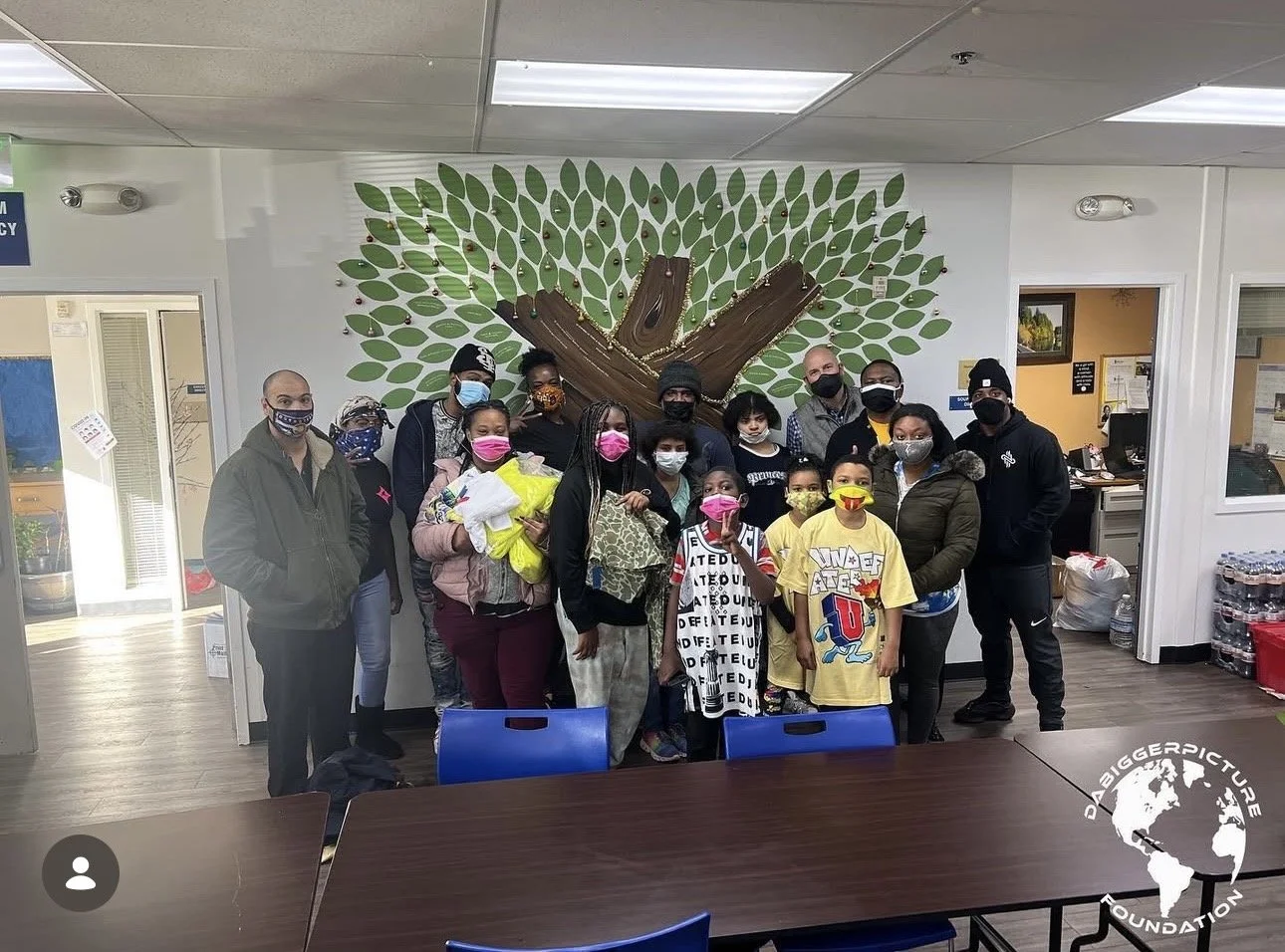 Group of adults and children wearing masks standing inside a building in front of a wall decorated with a large tree mural.