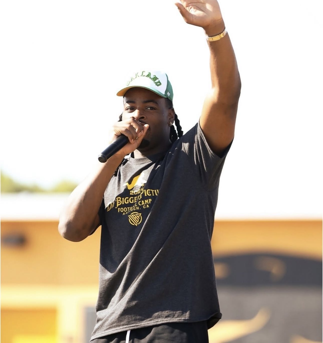 A young man with braided hair wearing a baseball cap, black T-shirt, and gold wristwatch, speaking into a microphone at an outdoor event.