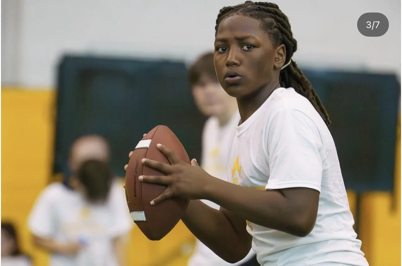 A young girl with braided hair holding a football, looking towards the camera, in a gym setting with children in white shirts in the background.
