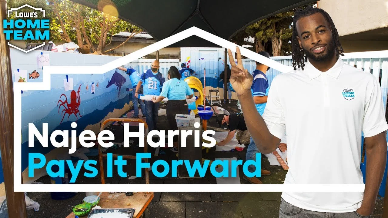 A man wearing a white Lowe's Home Team polo shirt making a peace sign while standing outdoors in front of a mural with people working on undersea-themed art projects, such as a crab, fish, and dolphin, in a yard with trees and a white fence.