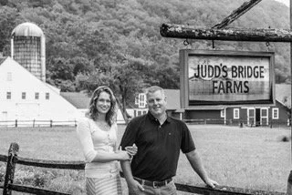 A smiling man and woman standing together in front of a sign that reads 'Judd's Bridge Farms' with farm buildings and trees in the background.