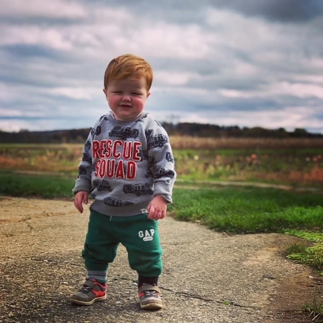 A young boy with red hair standing outdoors on a dirt path with fields and cloudy sky in the background. He is wearing a grey sweatshirt with 'Rescue Squad' written in red and dark pants.