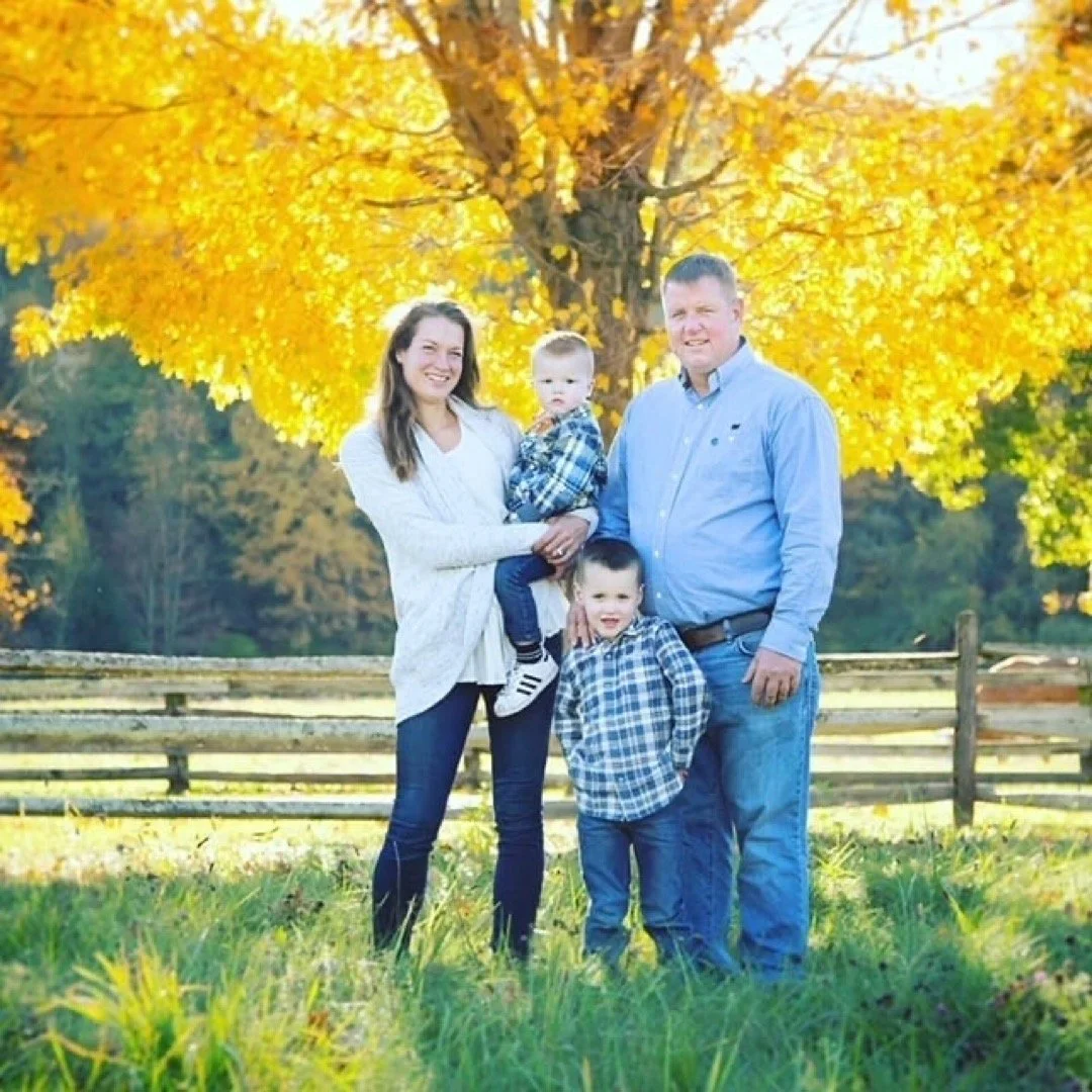 A family of four standing outdoors in front of a large tree with yellow autumn leaves. The mother holds a young boy, and the father stands beside her with a young boy in front of him. They are all smiling, dressed in casual clothes, and surrounded by green grass.