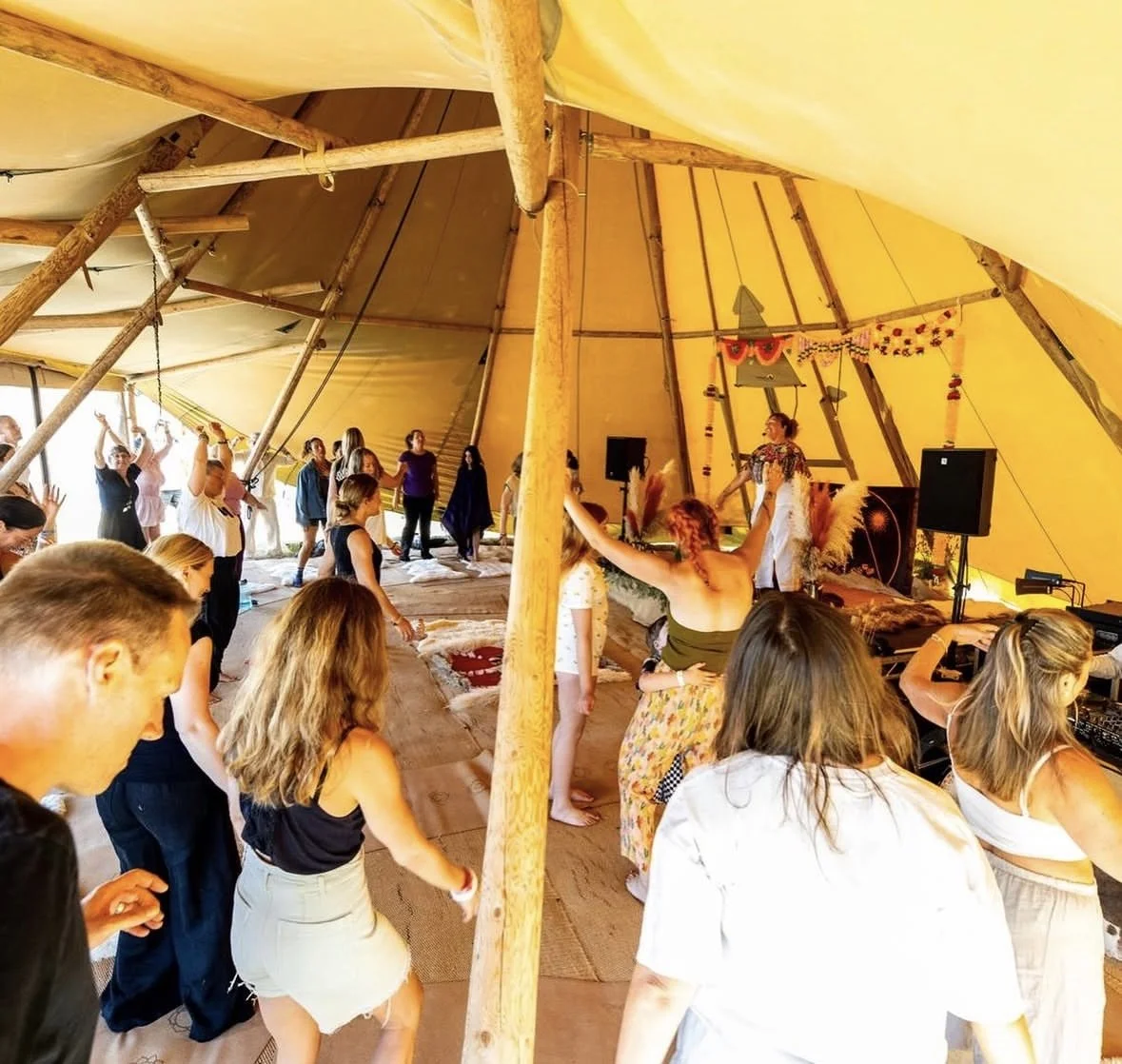 People gathered inside a yellow tipi tent, participating in a dance or ceremony, with a woman standing at the front and by speakers, decorations, and wooden support beams visible.