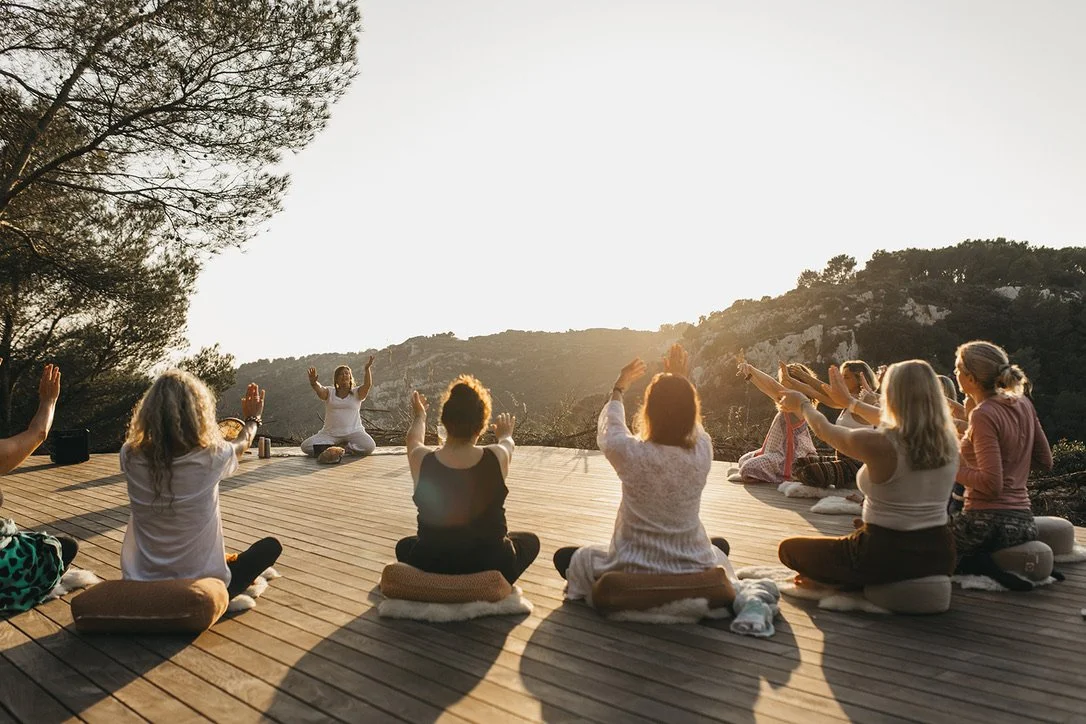 Group of people practicing yoga outdoors on a wooden deck during sunset, sitting on cushions with arms raised, surrounded by trees and hills.