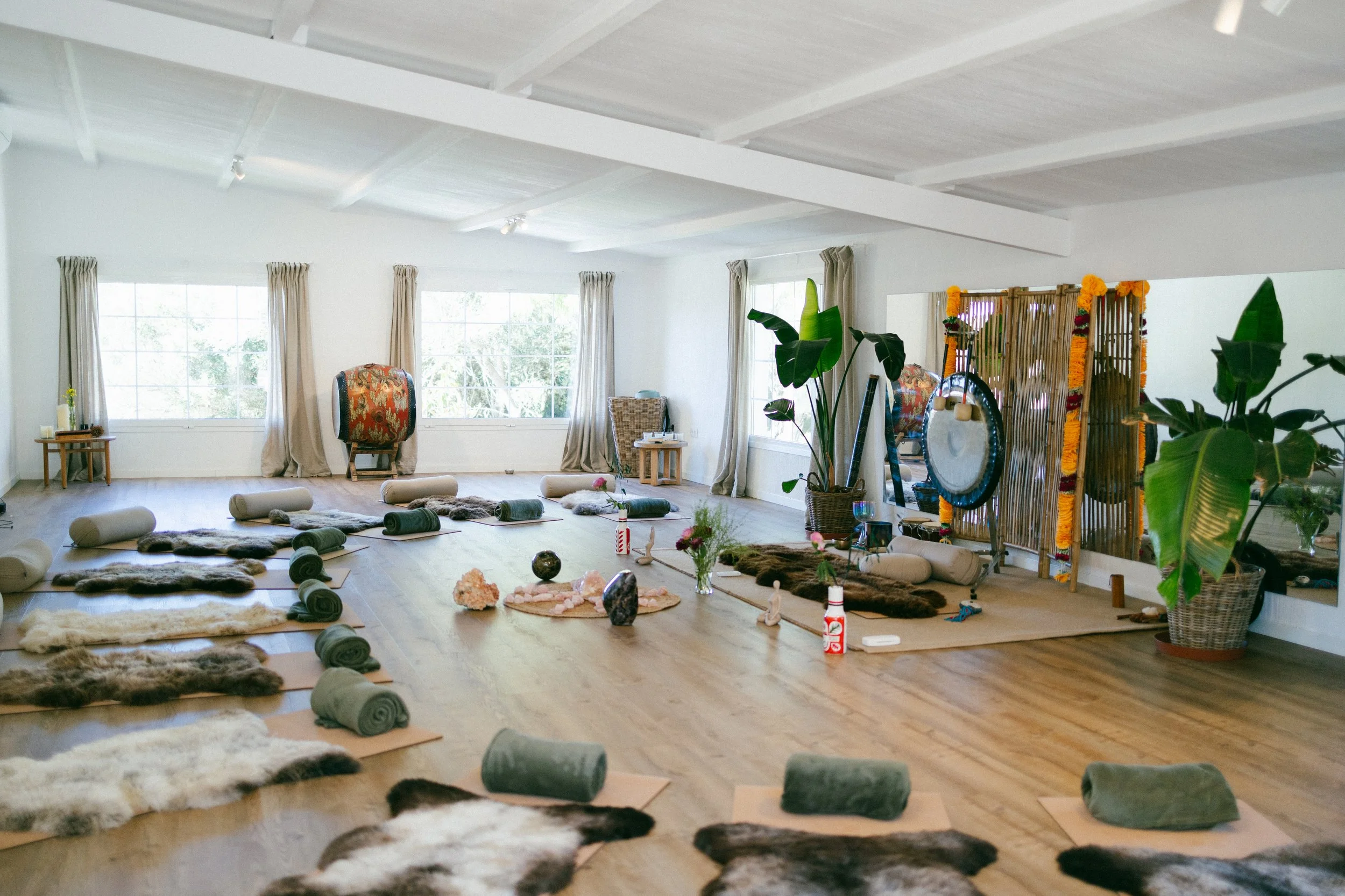 A spacious room prepared for a yoga or meditation class with mats, bolsters, and animal hide rugs arranged on the wooden floor. The room is decorated with plants, a folding screen with marigold flowers, and has large windows with curtains letting in 