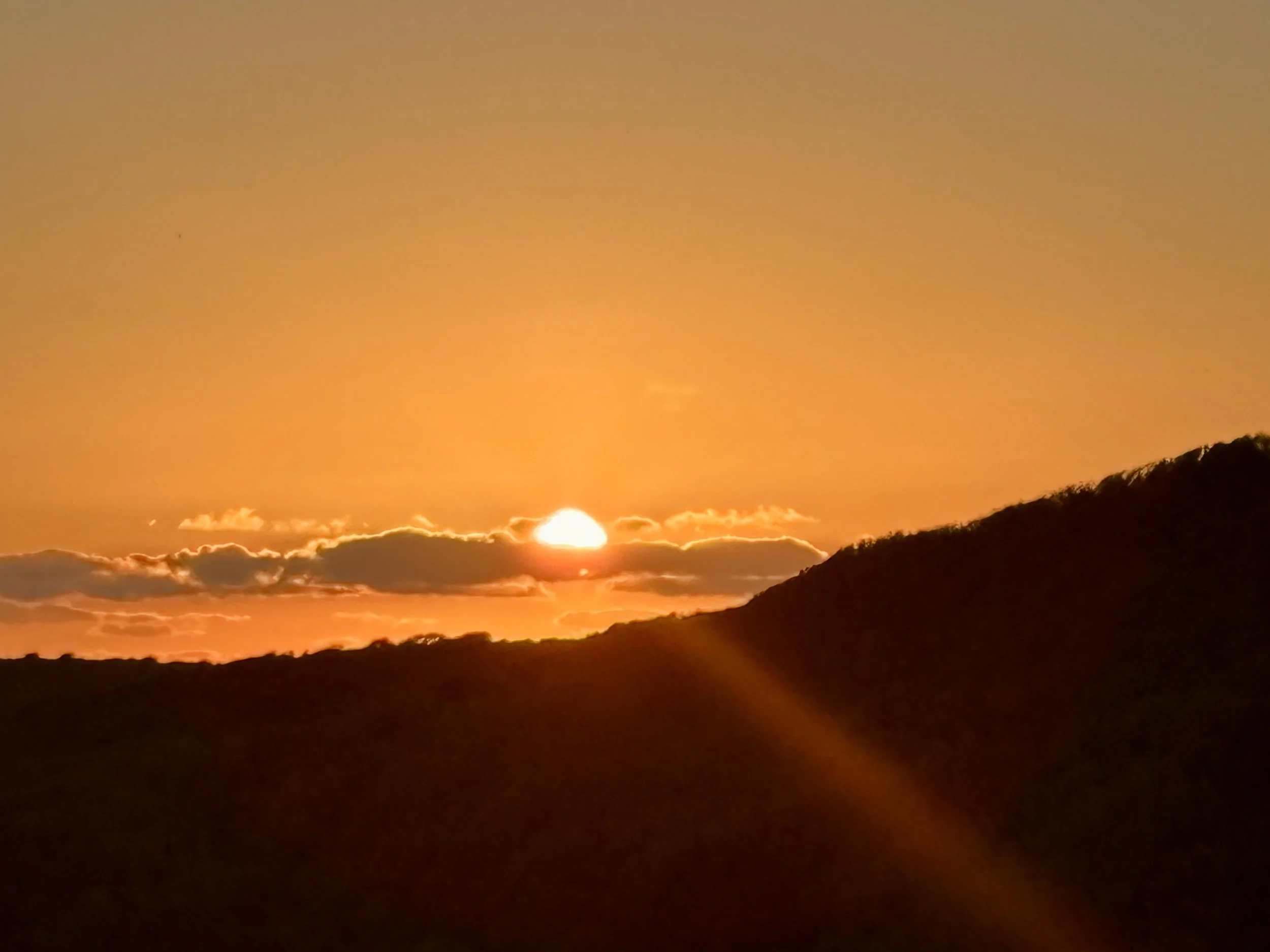 Sun setting over a landscape with silhouetted hills and clouds, creating an orange glow in the sky.