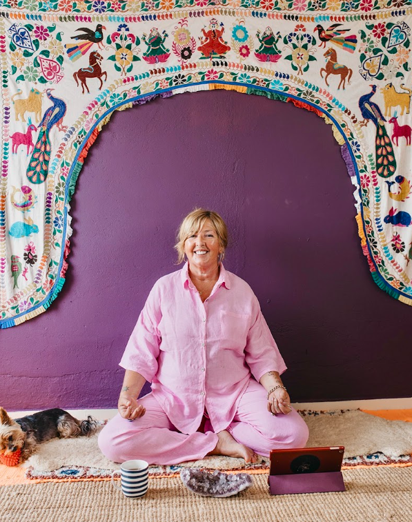 A woman sitting cross-legged on a patterned rug in front of a purple wall, smiling at the camera, with a dog lying nearby, a striped mug, an electronic device on a stand, and colorful embroidered curtains with animals and floral patterns hanging above.