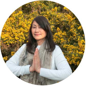 A woman stands outdoors with her hands pressed together in a prayer-like gesture, surrounded by yellow flowering bushes.