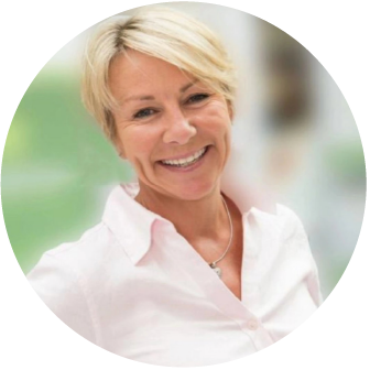 A smiling older woman with short blonde hair wearing a white blouse, indoors with a blurred background.