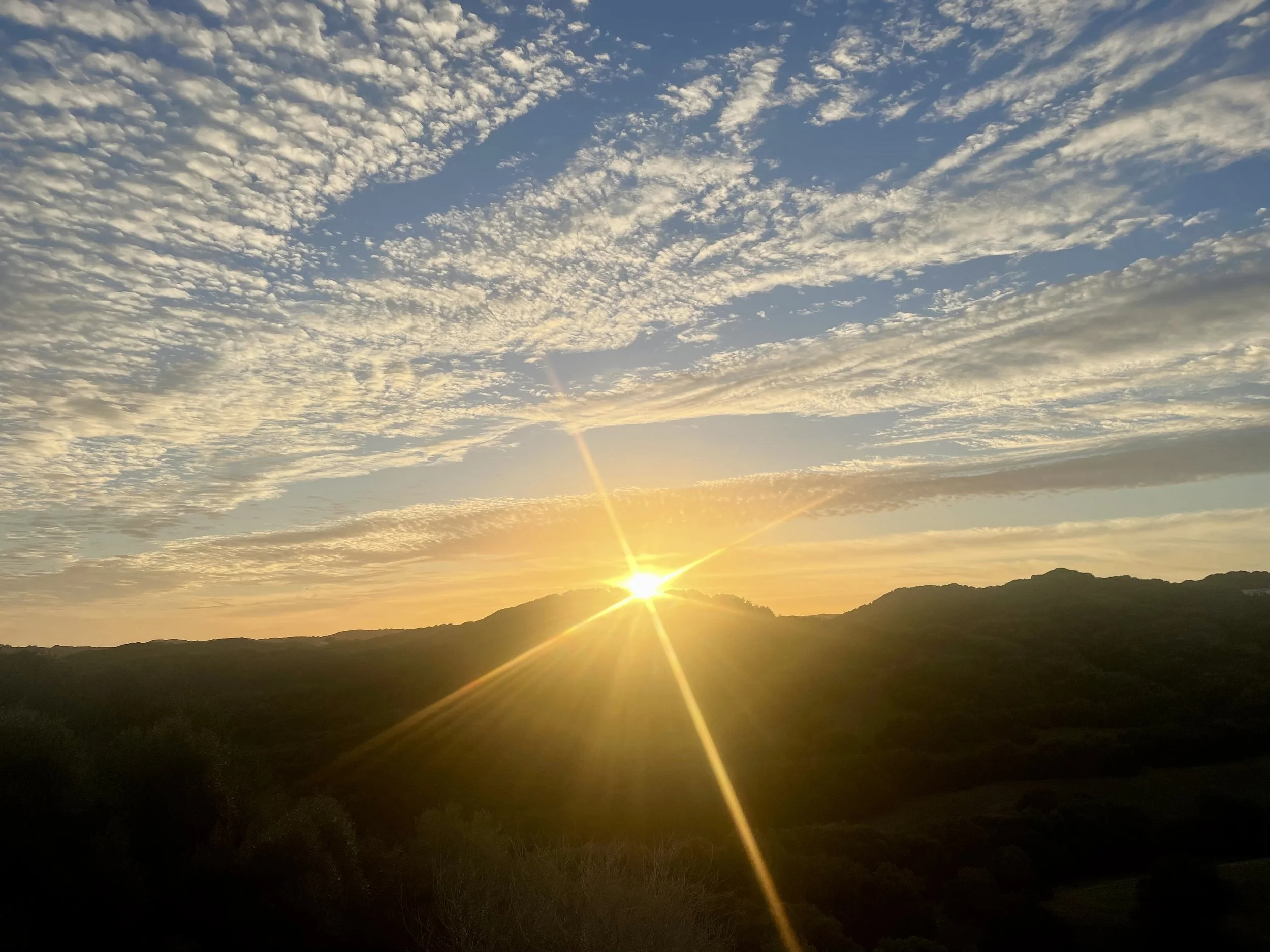 A sunset over a landscape with rolling hills in the distance and a sky filled with scattered clouds.