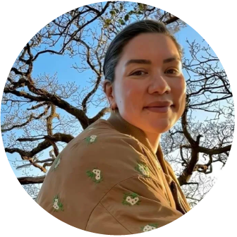Woman with dark hair pulled back, wearing a tan jacket with small embroidered designs, standing outdoors with leafless trees and a blue sky in the background.
