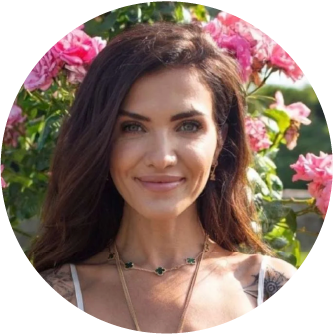 A woman with long dark hair smiling outdoors, with pink flowers in the background.