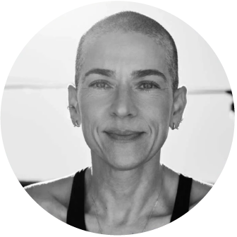 Close-up black and white photo of a woman with a shaved head, smiling slightly, wearing earrings and a tank top.