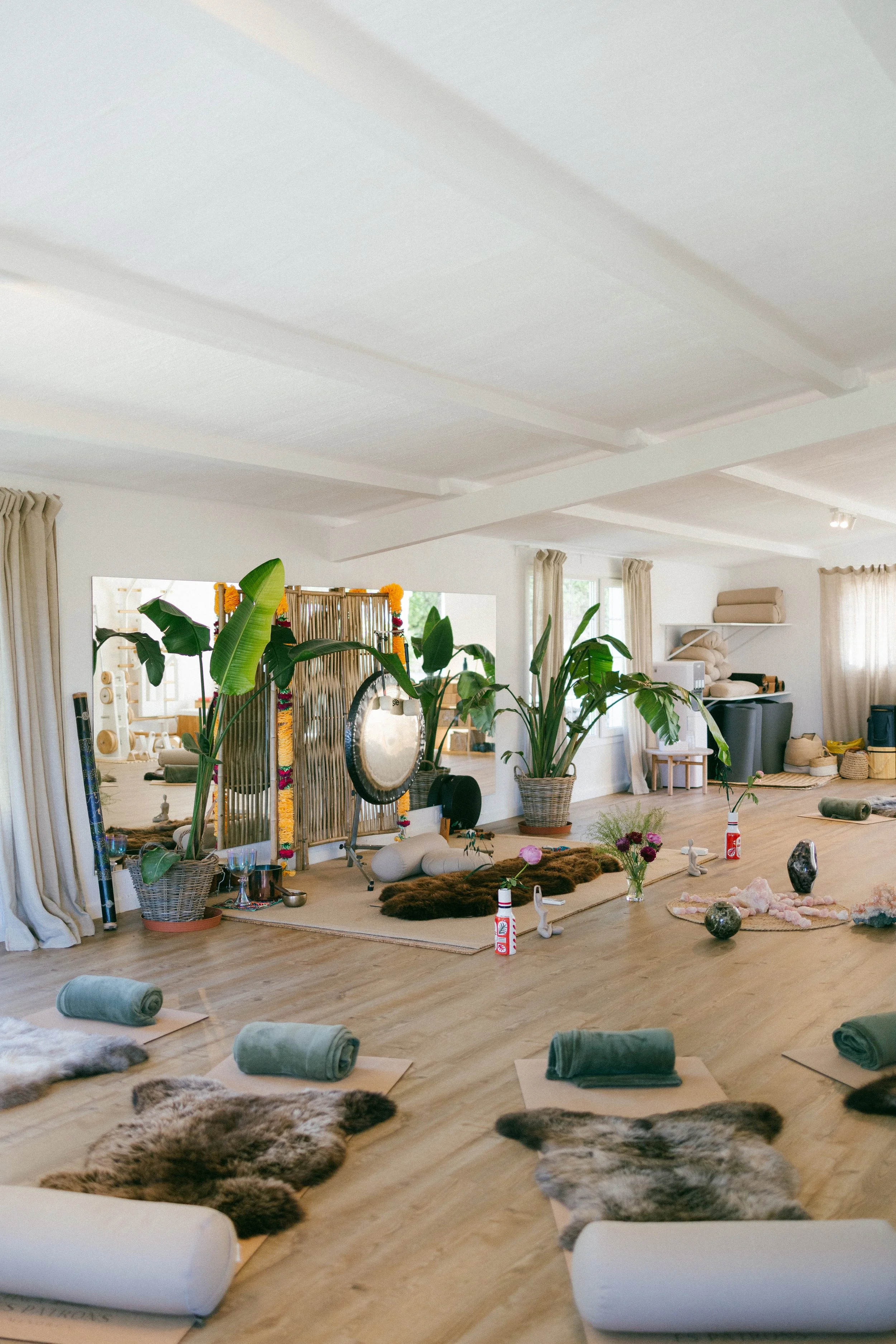 Interior of a bright living space set up for a yoga or meditation class with mats, rolled towels, sheepskin rugs, and decorative objects, along with large green potted plants and natural light from windows.