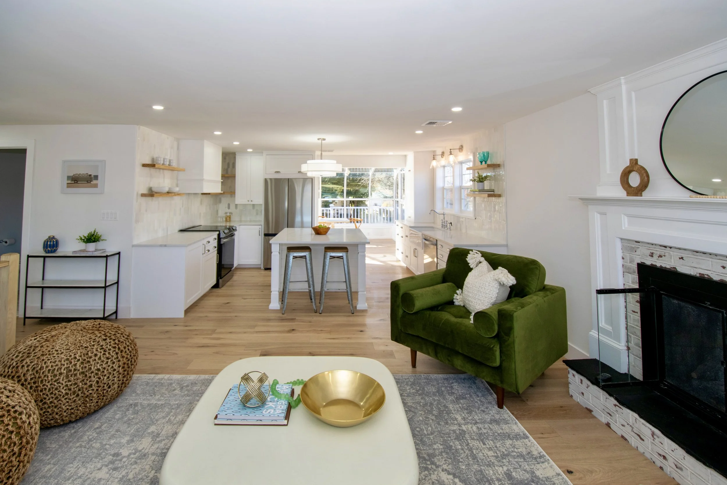 Open-concept living room and kitchen with white walls, wooden flooring, green armchair, fireplace, and large windows, decorated with modern and rustic accents.