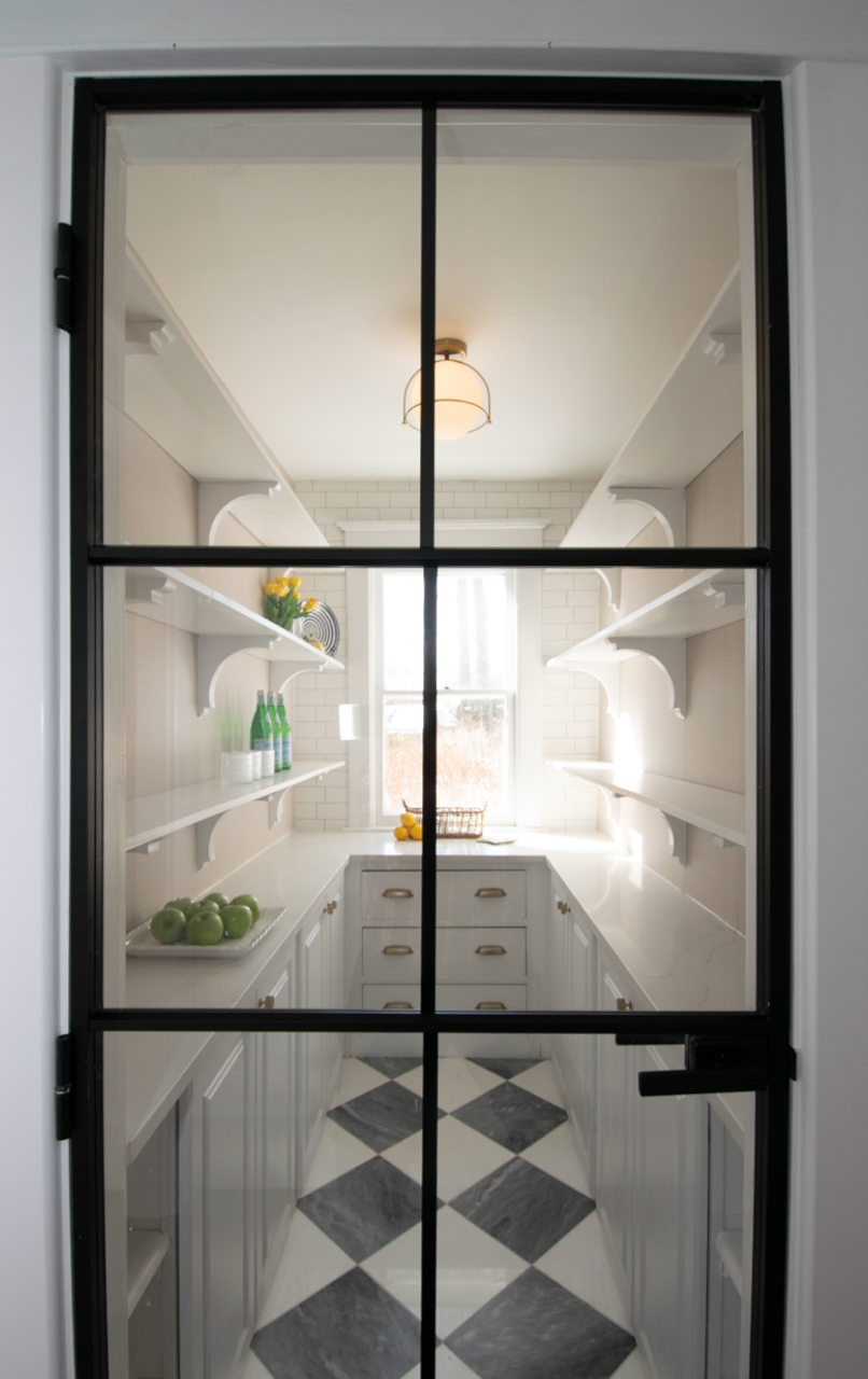 View through a glass-paneled door into a small, modern kitchen with white cabinets, open shelves, a window, and a black-and-white checkered floor.