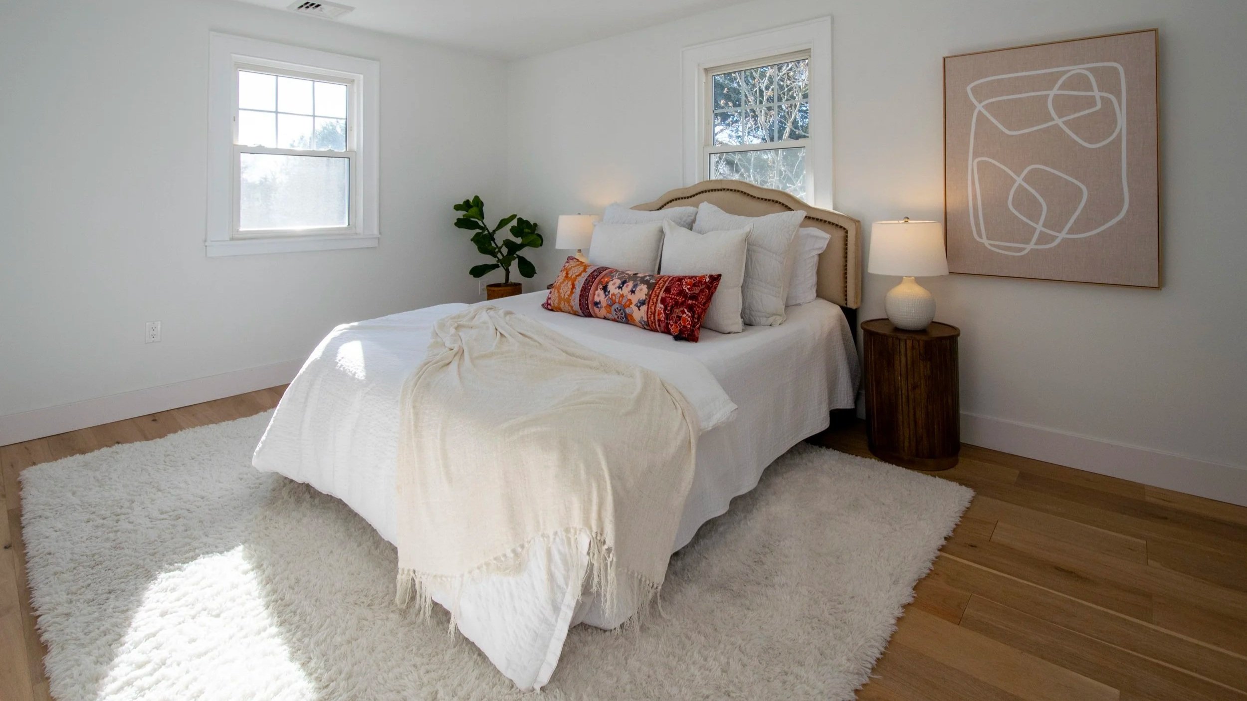 A bedroom with a white bed, multiple pillows, a rolled colorful decorative pillow, a wooden nightstand with a lamp, a potted plant, and wall art, with two windows letting in natural light.