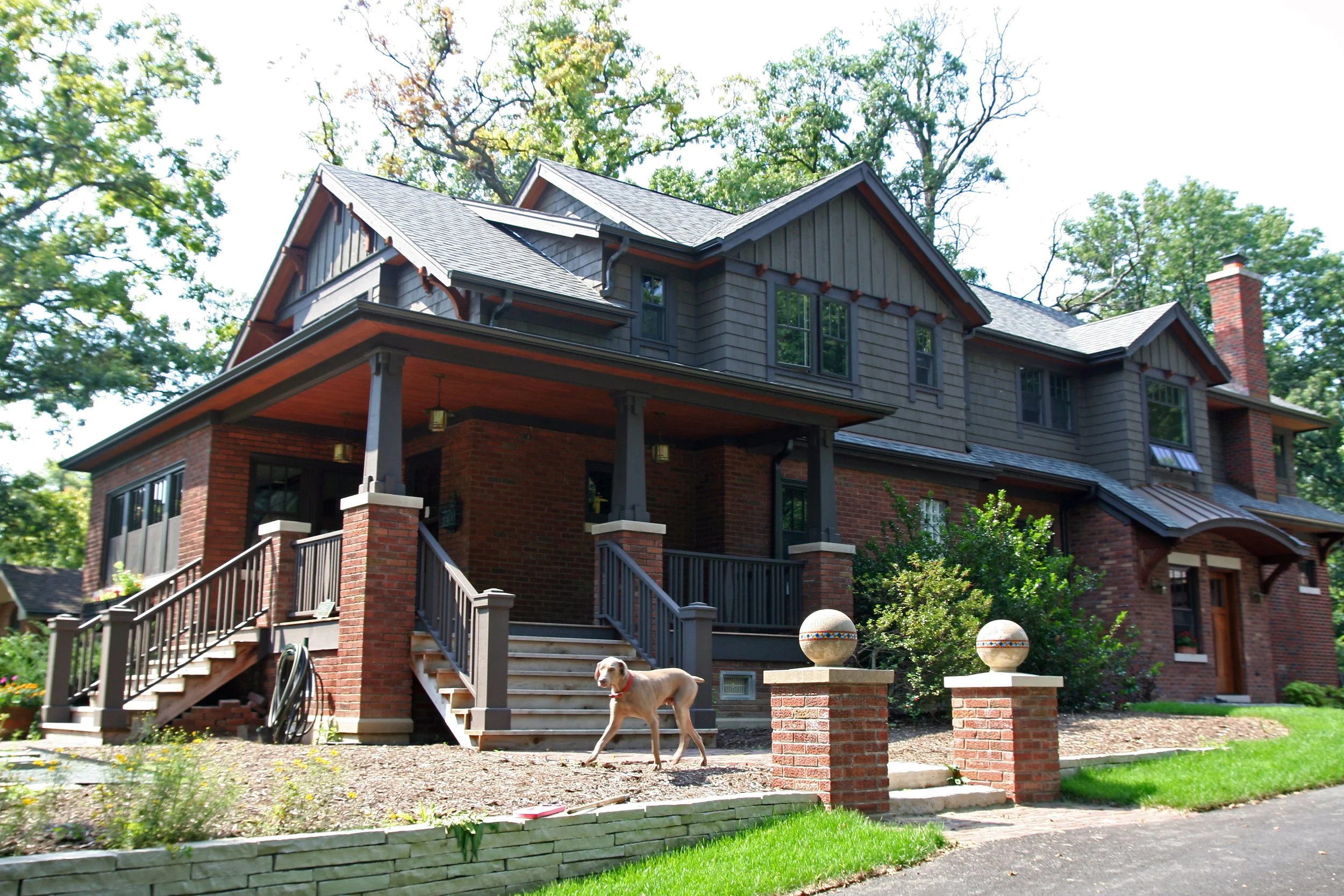 A large, multi-story house with a brick and gray wooden exterior, front porch, steps, and a dog walking on the yard in front.